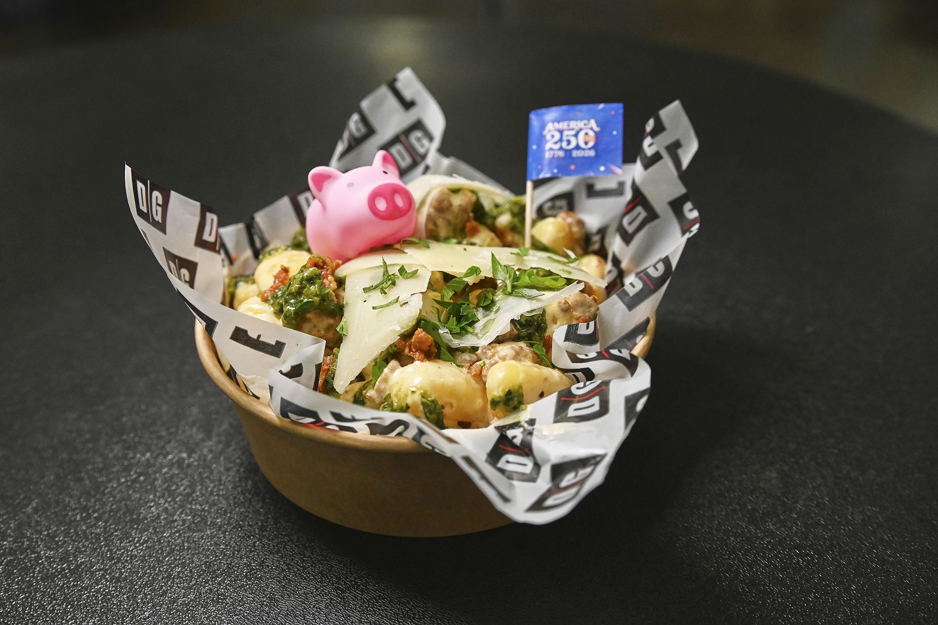 A bowl of loaded savory dish with potatoes, greens, and cheese shavings, wrapped in branded parchment, topped by a pink pig figurine and a blue "America 250" flag.