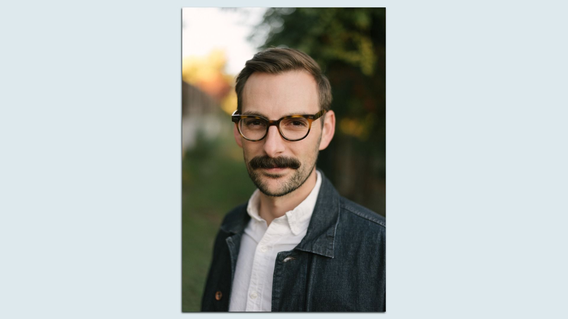 Portrait of a man with dark hair and mustache wearing glasses, a white shirt, and a dark jacket, standing outdoors with greenery and soft sunlight in the background.