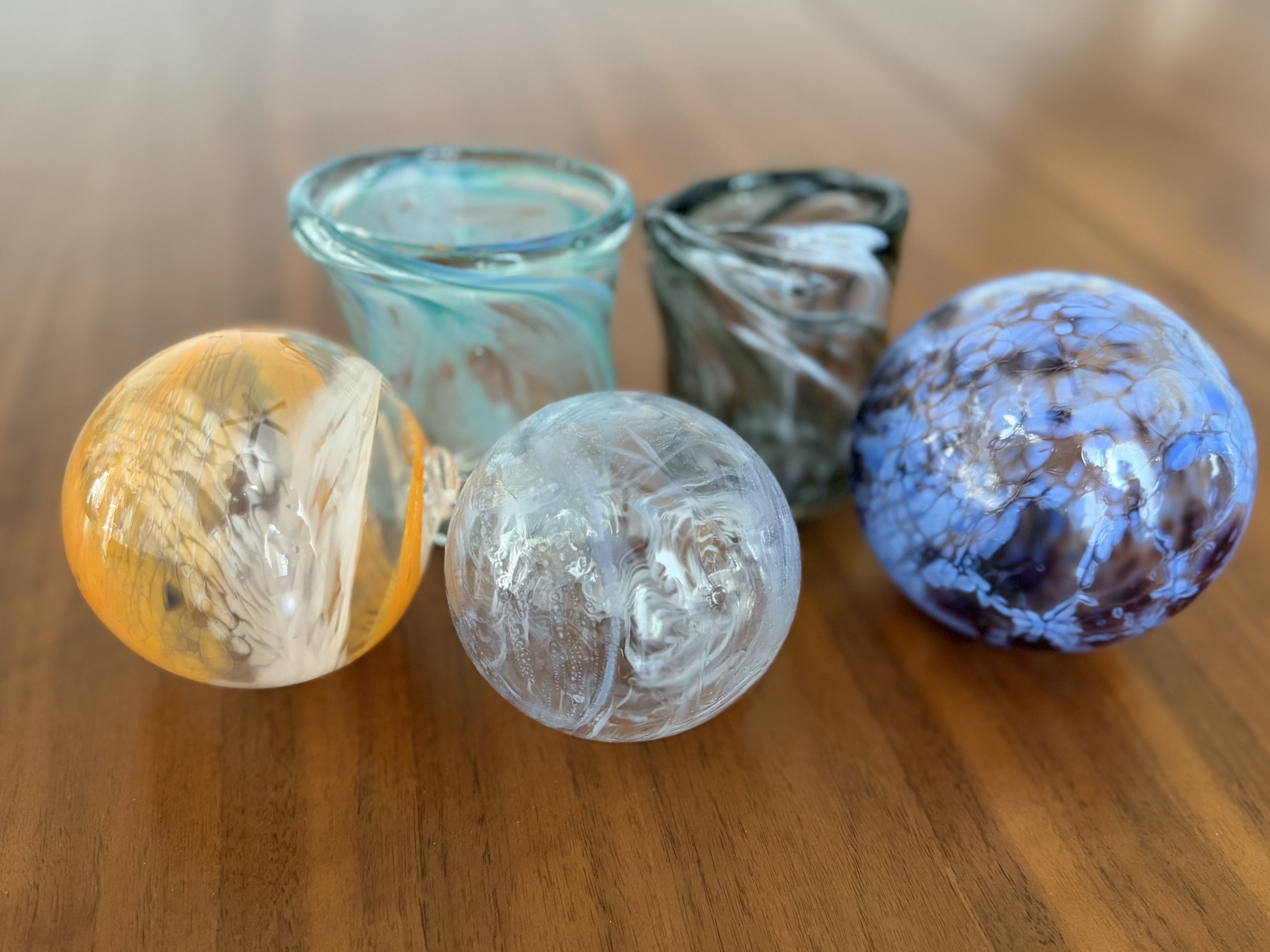 Close-up of colorful marbled glass spheres and turquoise glass cups on a warm wooden table; blue, teal, and orange swirls create a glass-art still life.