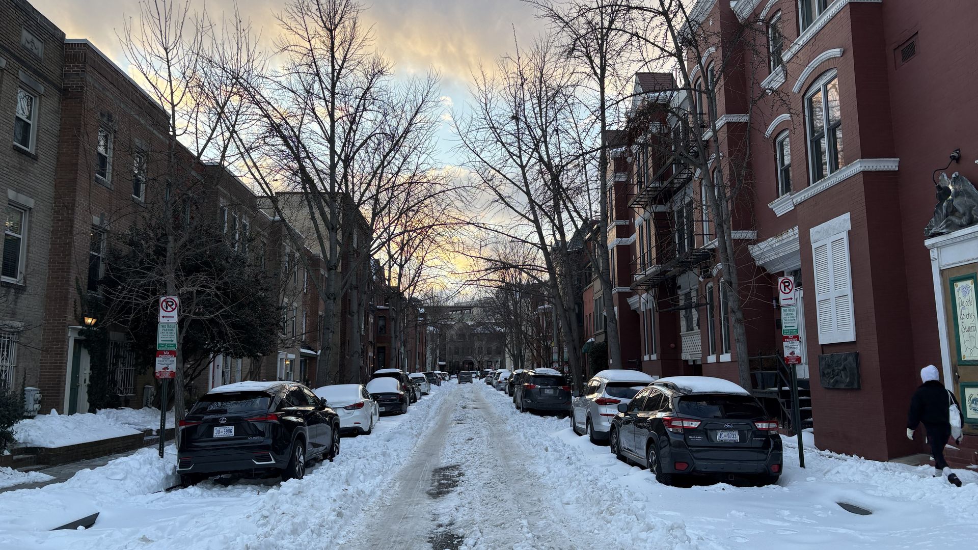 Snow-covered street with parked cars lined by brick buildings and leafless trees under a cloudy sky at sunset. A person wearing a white hood walks on the right sidewalk.