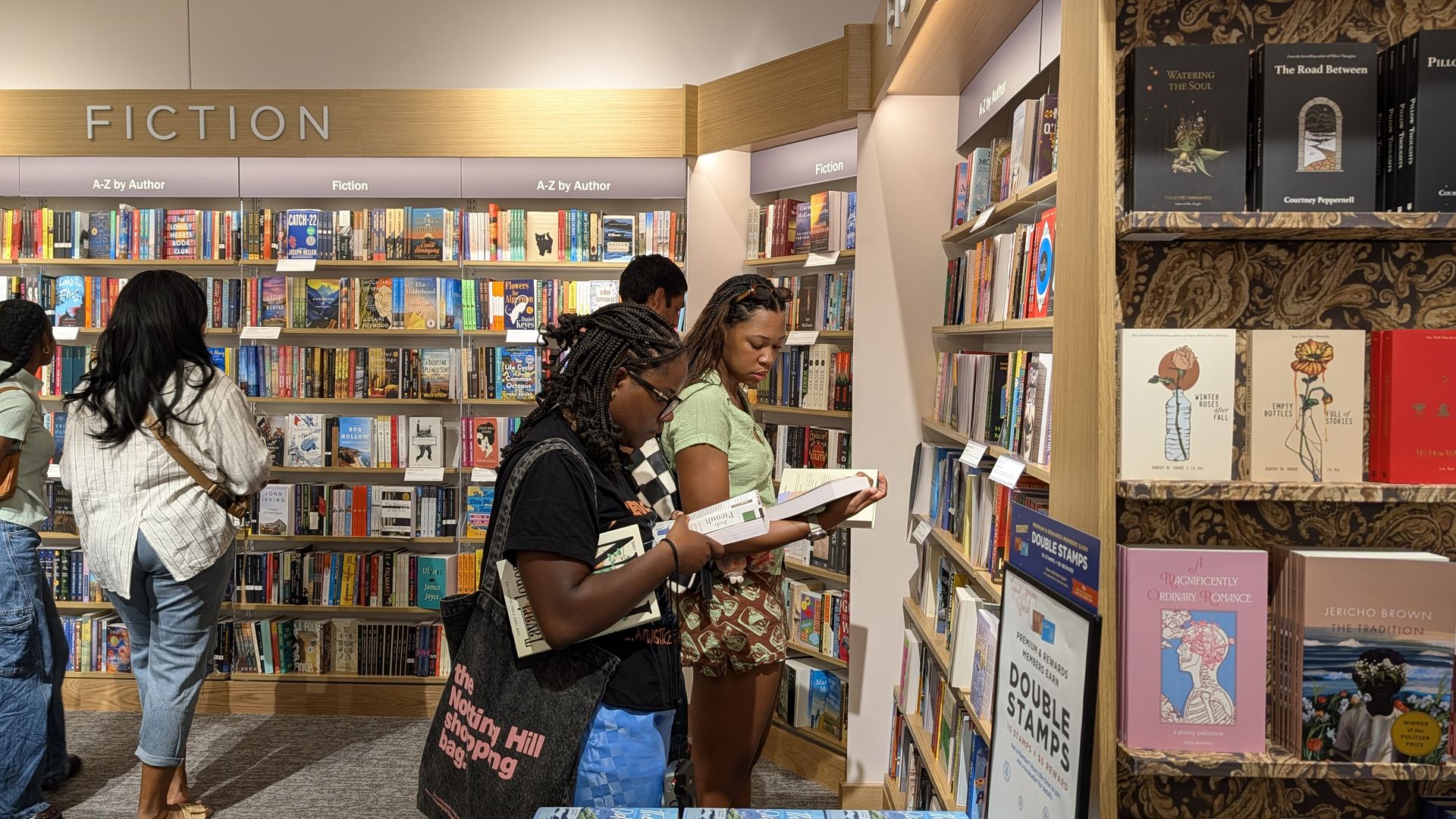 Inside a bookstore's fiction section, wooden shelves brim with colorful novels. Signs read FICTION and A-Z by Author as three women browse and read amid warm lighting.