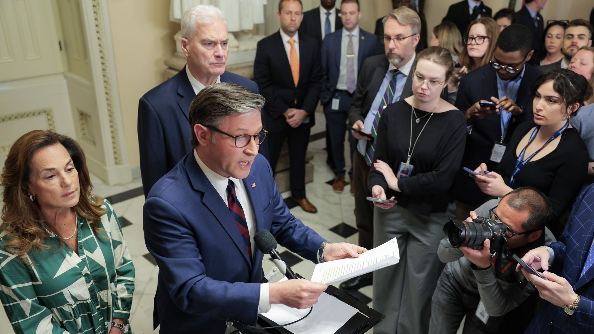A man in a blue suit speaks at a lectern with papers as reporters and photographers crowd a government building hallway; a woman in a green patterned dress stands nearby.