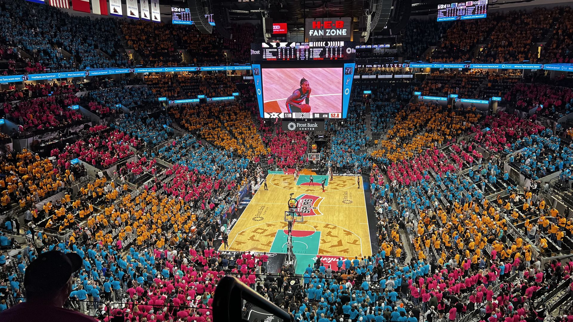Panoramic view of a packed basketball arena with fans in blue, pink, and yellow sections. A big center scoreboard shows a player on court; banners, flags, and a bright "H-E-B Fan Zone" sign hangs above.