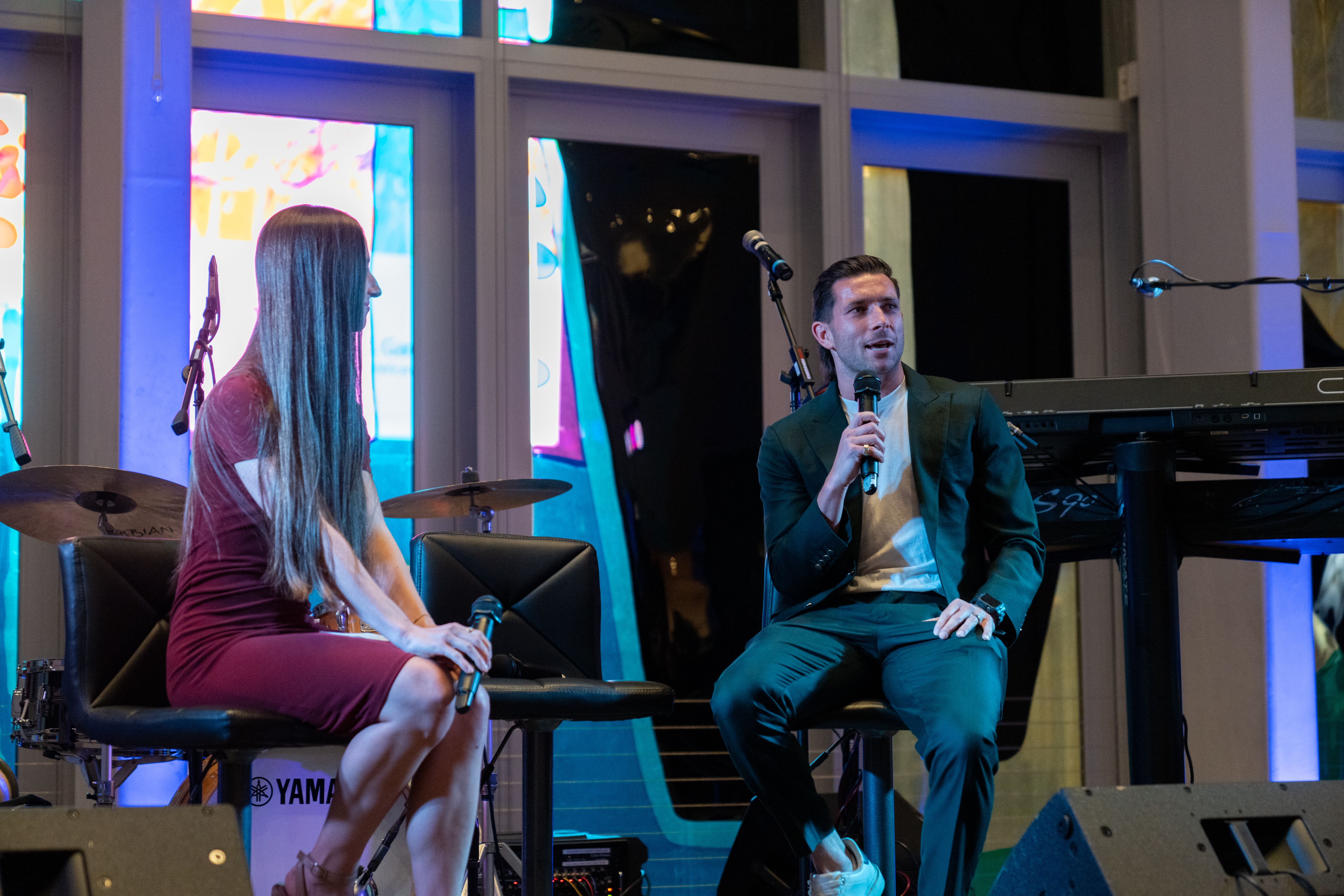 A man in a dark suit and white sneakers speaking into a microphone while sitting on a stool, and a woman in a maroon dress holding a microphone, sitting beside him on stage under blue lighting.