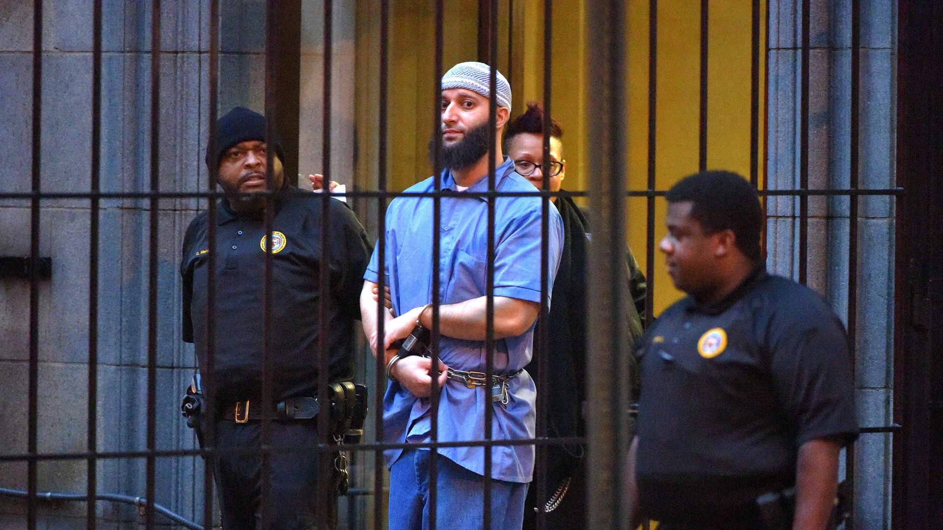 Photo of Adnan Syed in a blue prison jumpsuit as he's escorted by guards behind bars