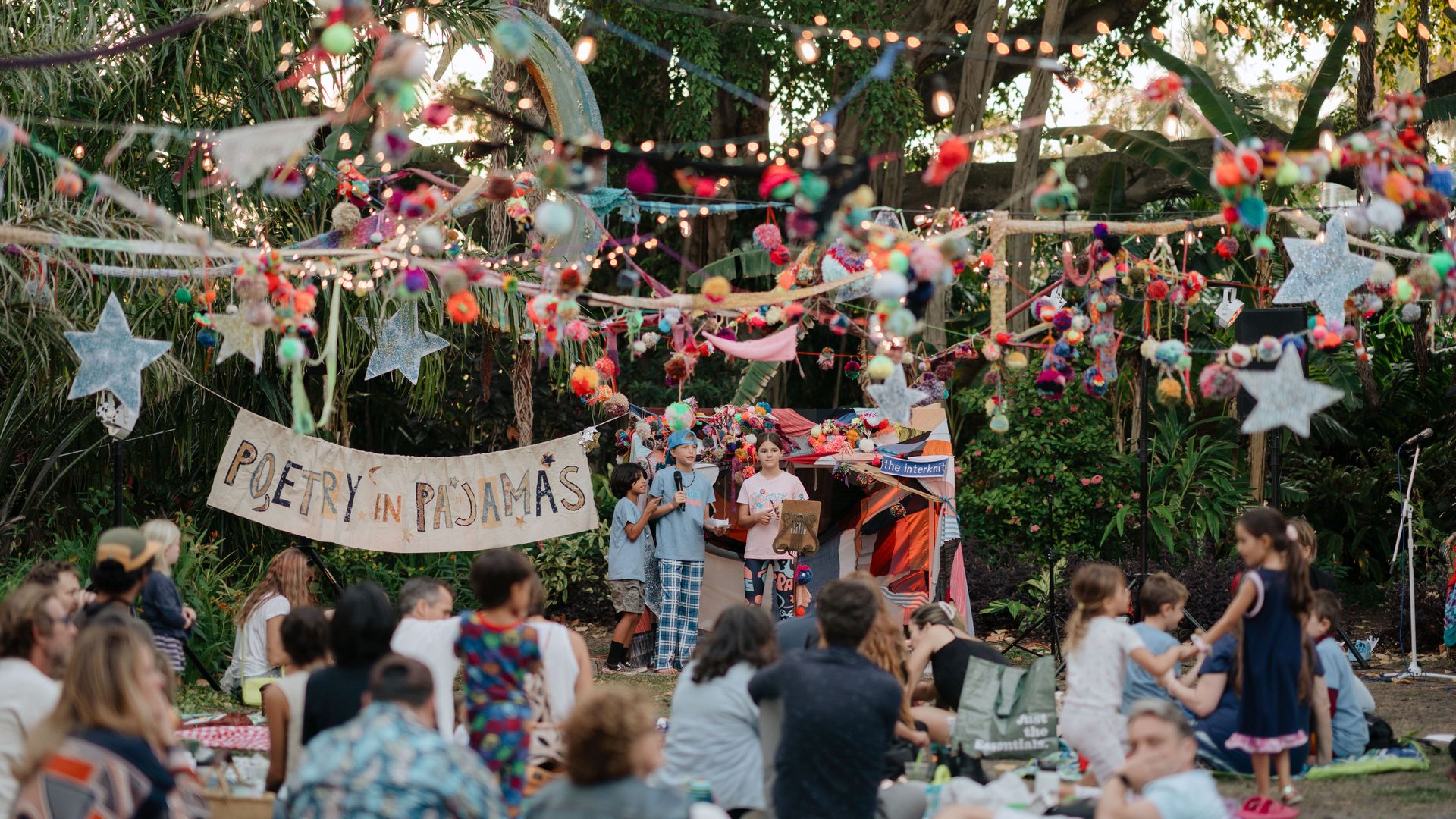 Outdoor garden festival with twinkling string lights and star garlands. Children perform on a small stage while a crowd sits on blankets.