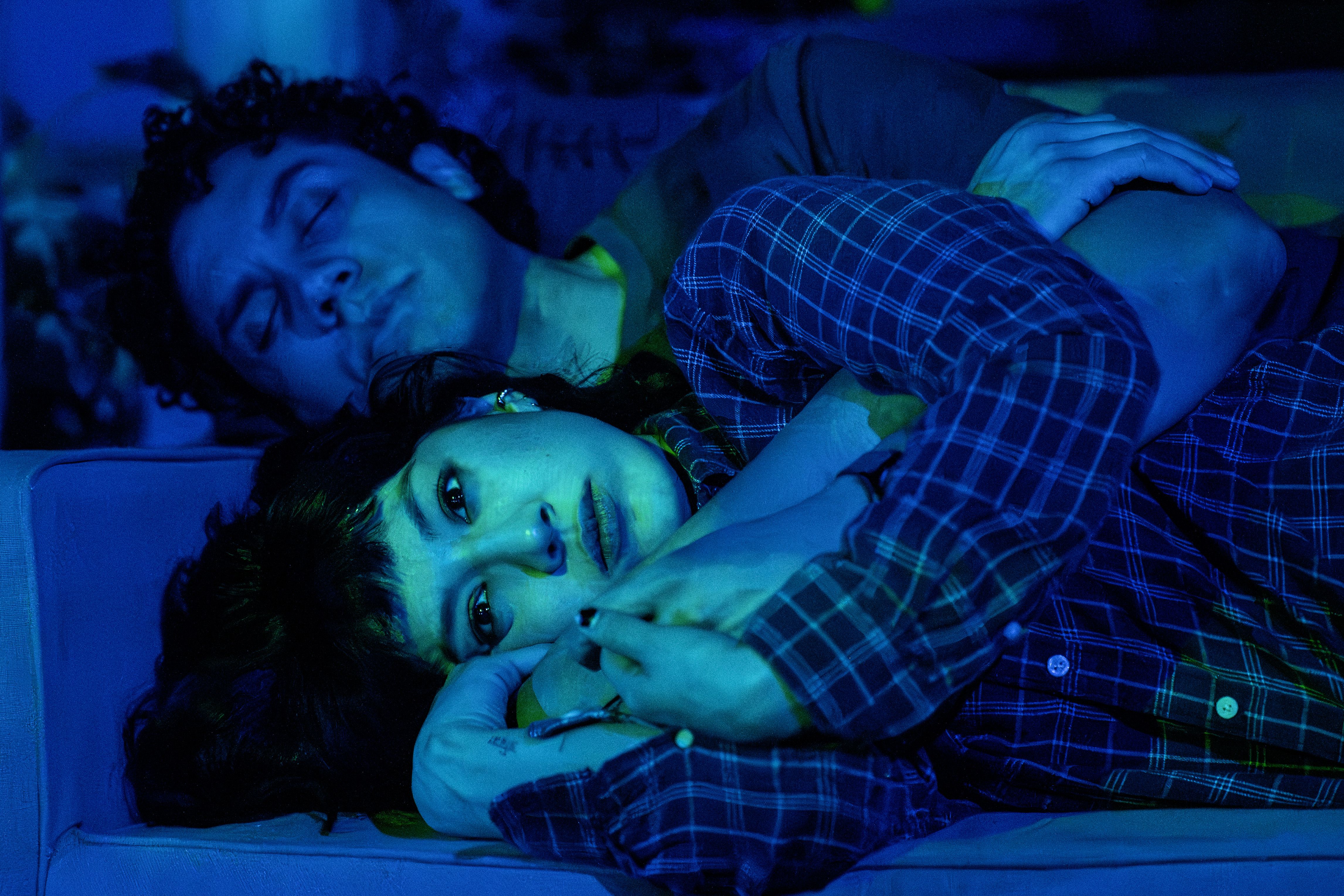 Two people lying on a couch under blue light; one has curly hair and is asleep, the other with straight hair and a plaid shirt looks thoughtful, holding a pen and resting their head on the other's arm.