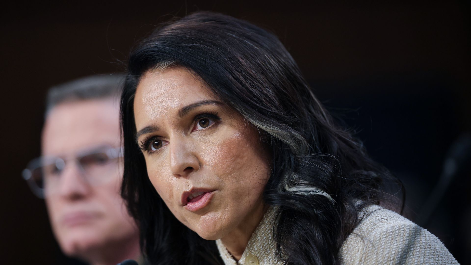 Tulsi Gabbard, who has dark hair with a lighter face-framing streak, leans forward and speaks into a microphone during a congressional hearing while wearing a beige jacket.