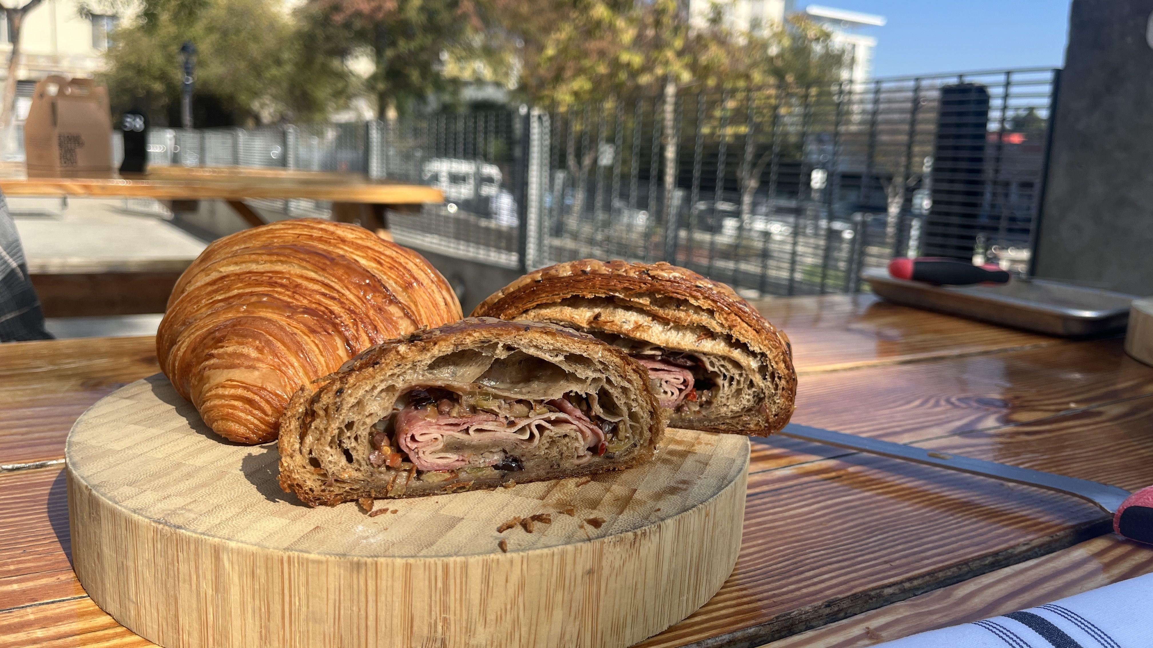 Two croissants on a round wooden board on an outdoor wooden table, one plain and one cut in half showing folded meat and vegetable filling, with trees and buildings in background.