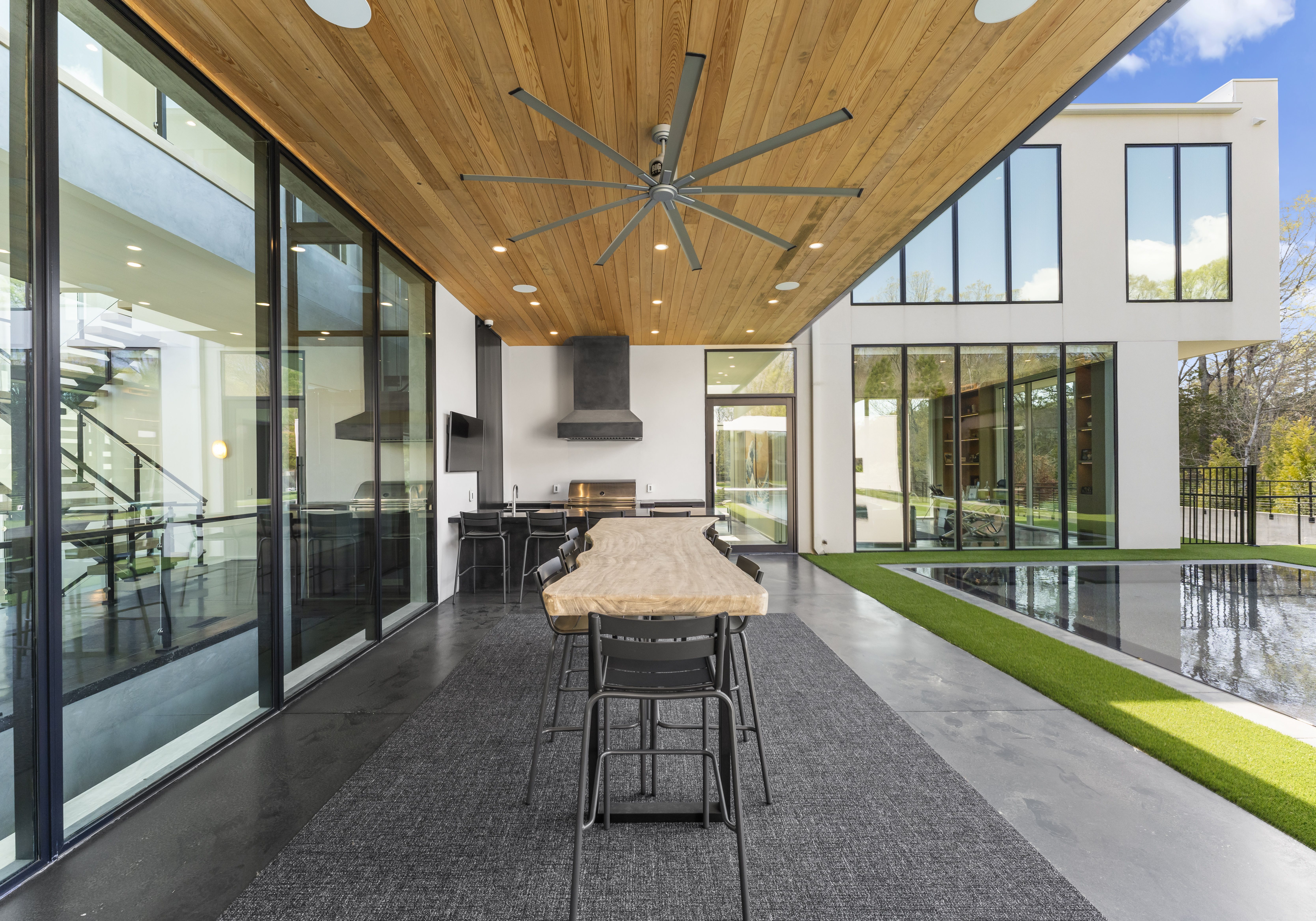 Modern outdoor dining area with a natural wood table, black metal chairs, gray rug, and ceiling fan under a wooden ceiling, next to large glass windows and a reflective pool.