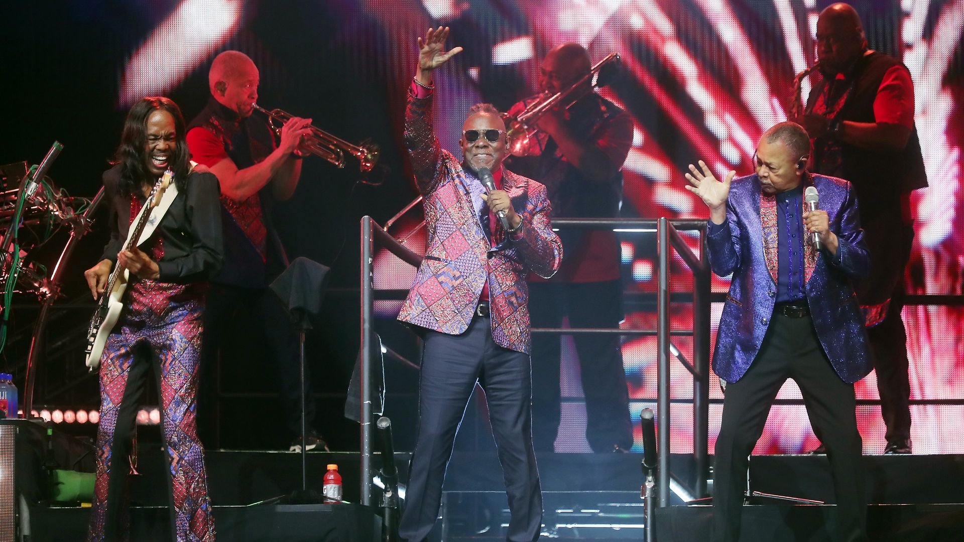 A photo showing three performers on a stage illuminated by lights with three background wind players behind them. Two of the main performers are wearing shiny jackets and holding microphones. The other is smiling and playing guitar with shiny pants.