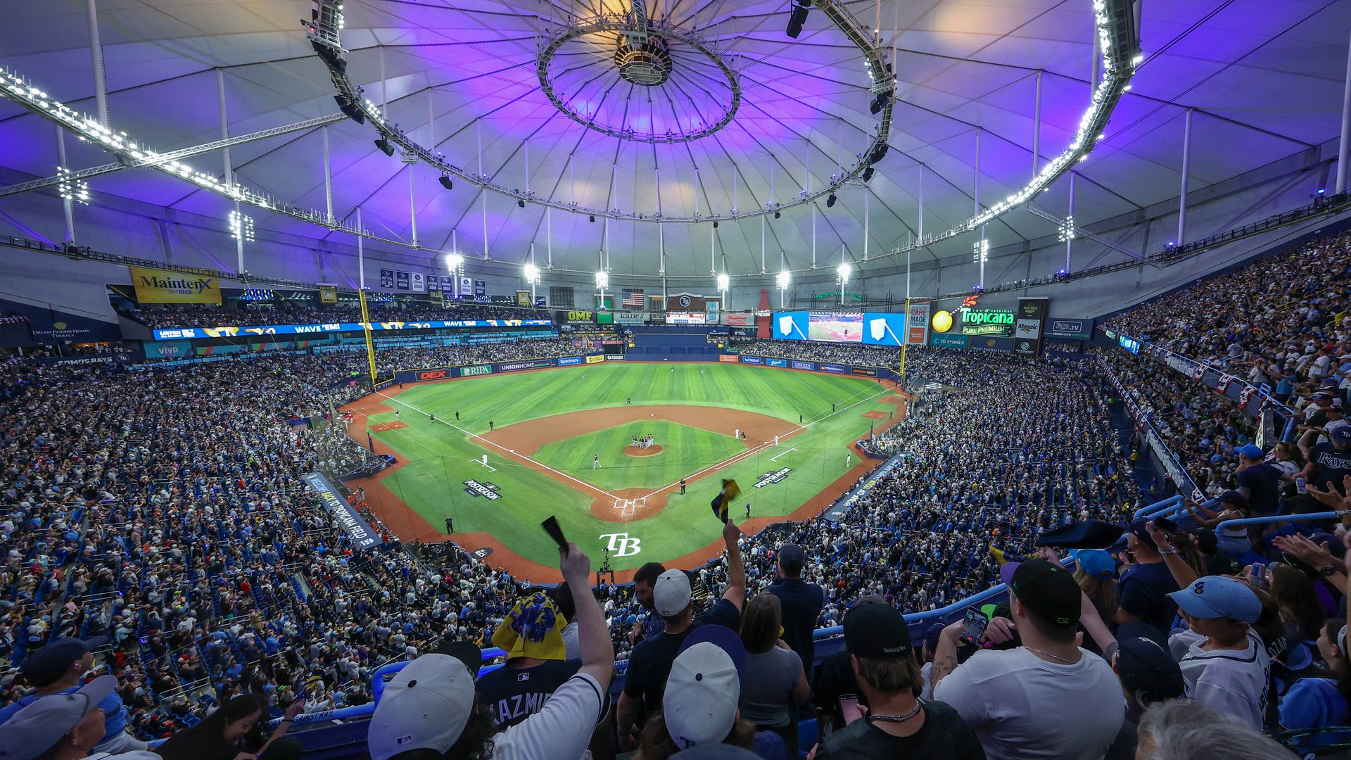 Panoramic view of a packed baseball stadium with a circular overhead rig casting purple lighting. Green field, players near home plate, and dense blue-clad fans filling the stands.