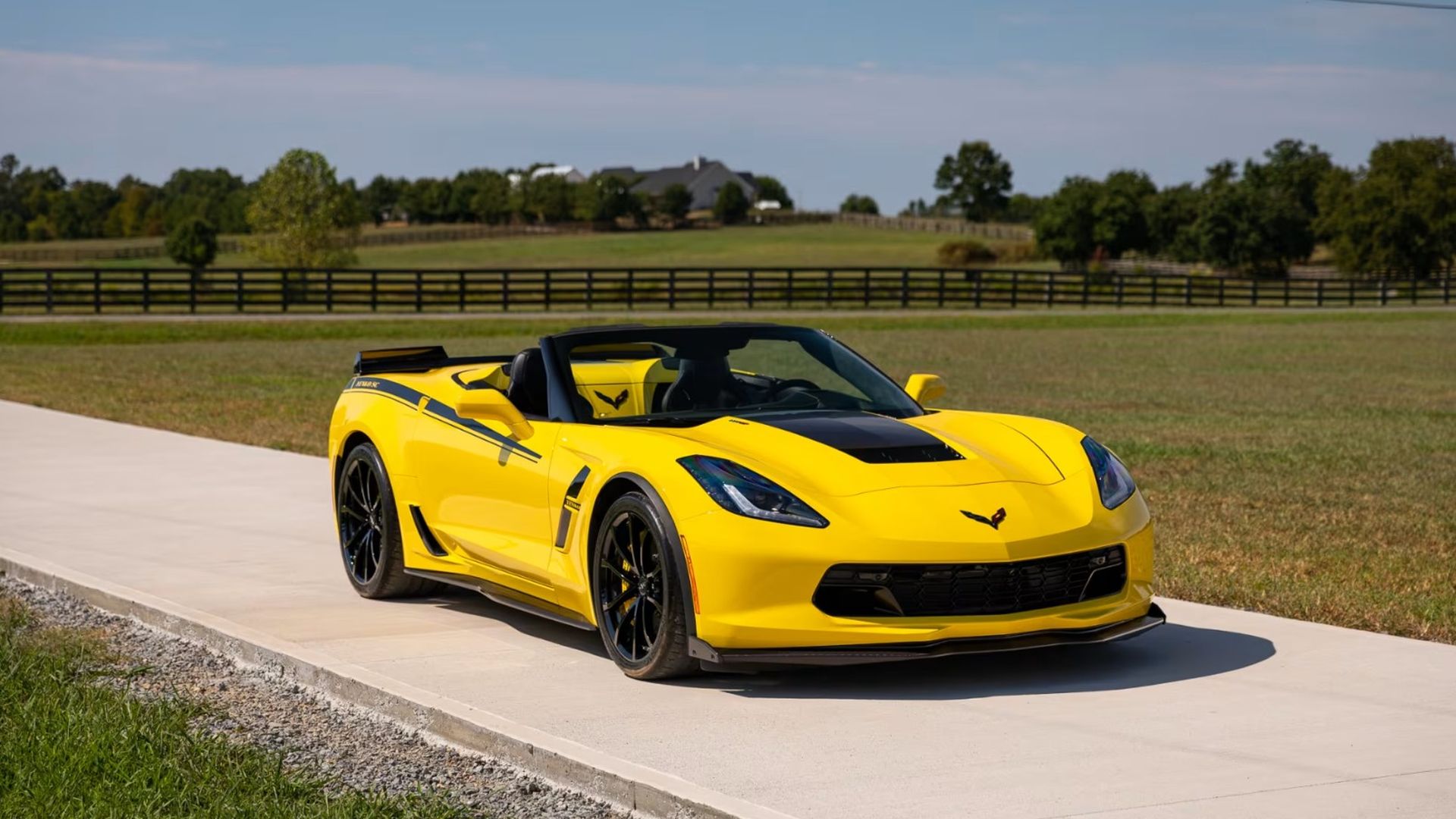Bright yellow convertible sports car with black accents parked on a concrete driveway, green fields and a fence in the background under a clear blue sky.