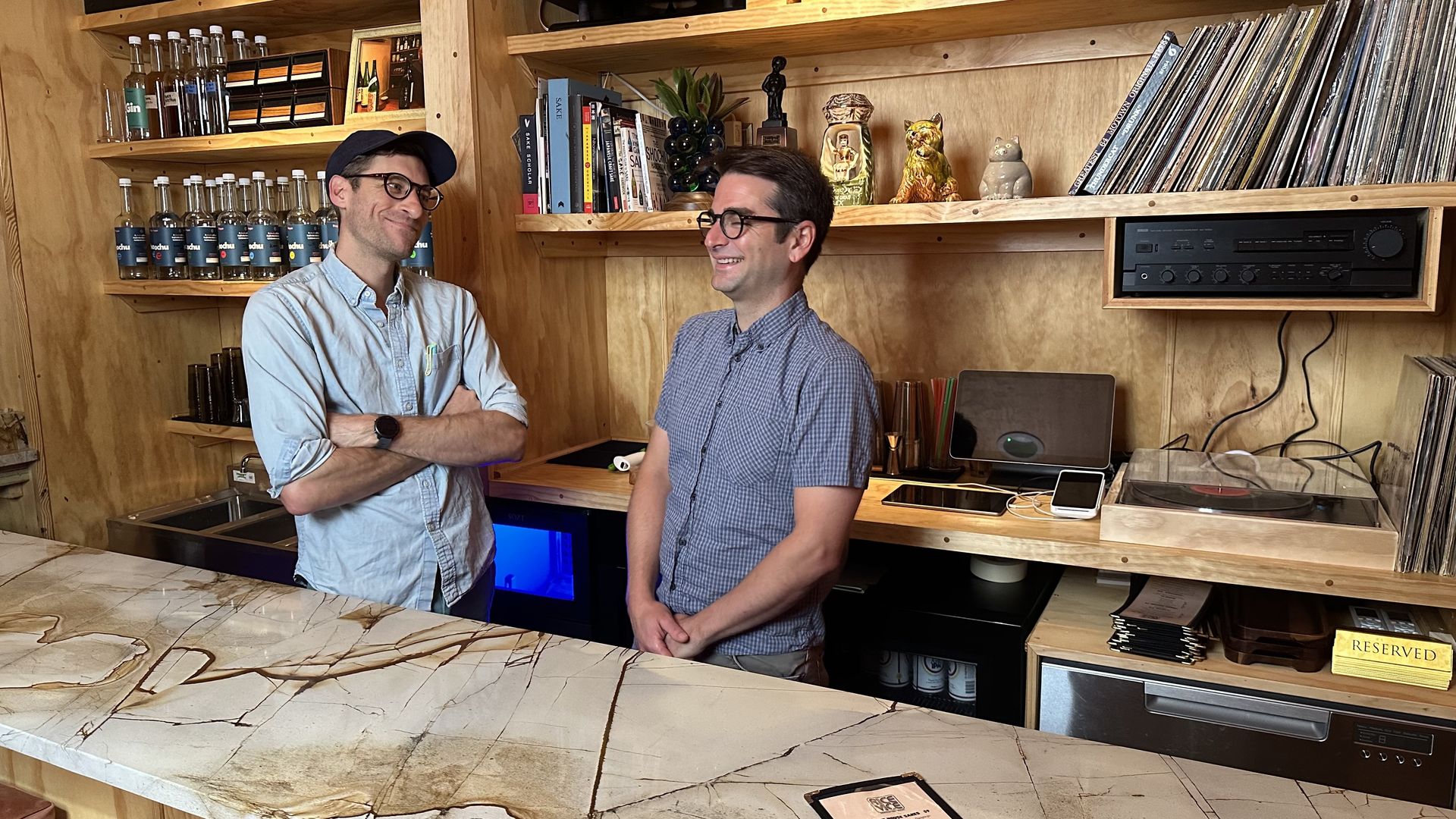 Two men stand behind a bar and laugh.