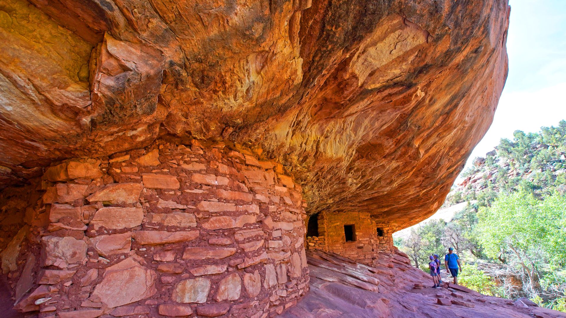 In this image, two hikers walk past a large clay and brick building