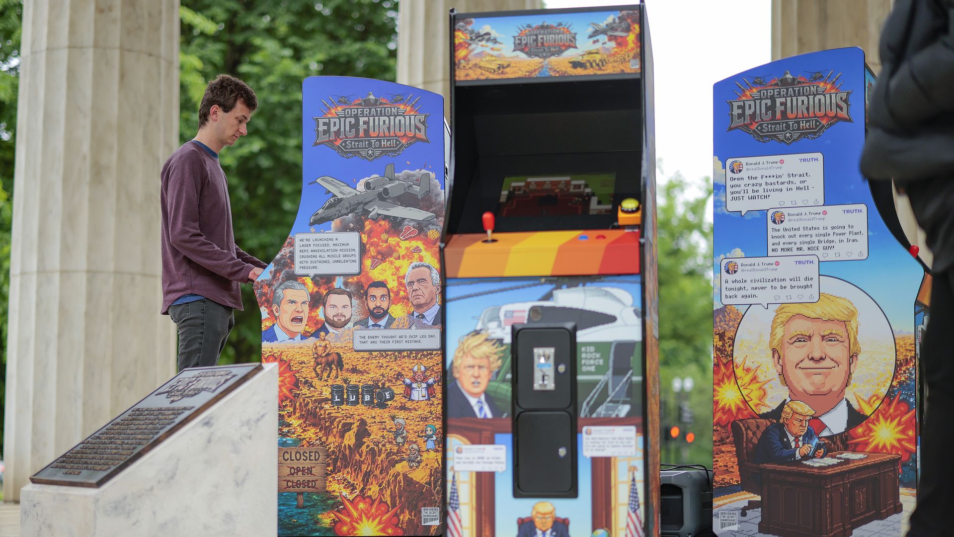 Young man in maroon sweater stands beside colorful arcade cabinet panels featuring warlike artwork and "Operation Epic Furious" branding; stone columns and green trees in background.