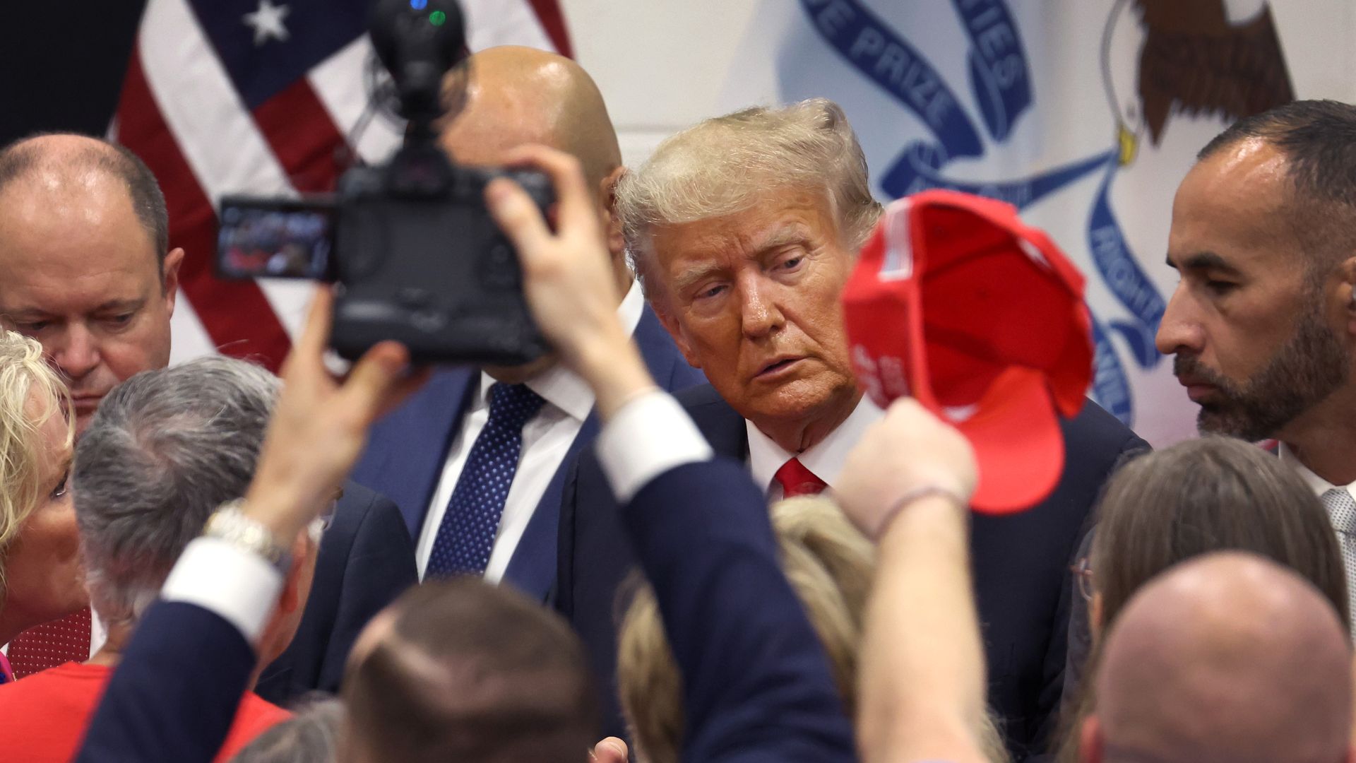 Former President Trump behind a crowd in Iowa.  A photographer holds up a camera and someone else waves a red hat. 