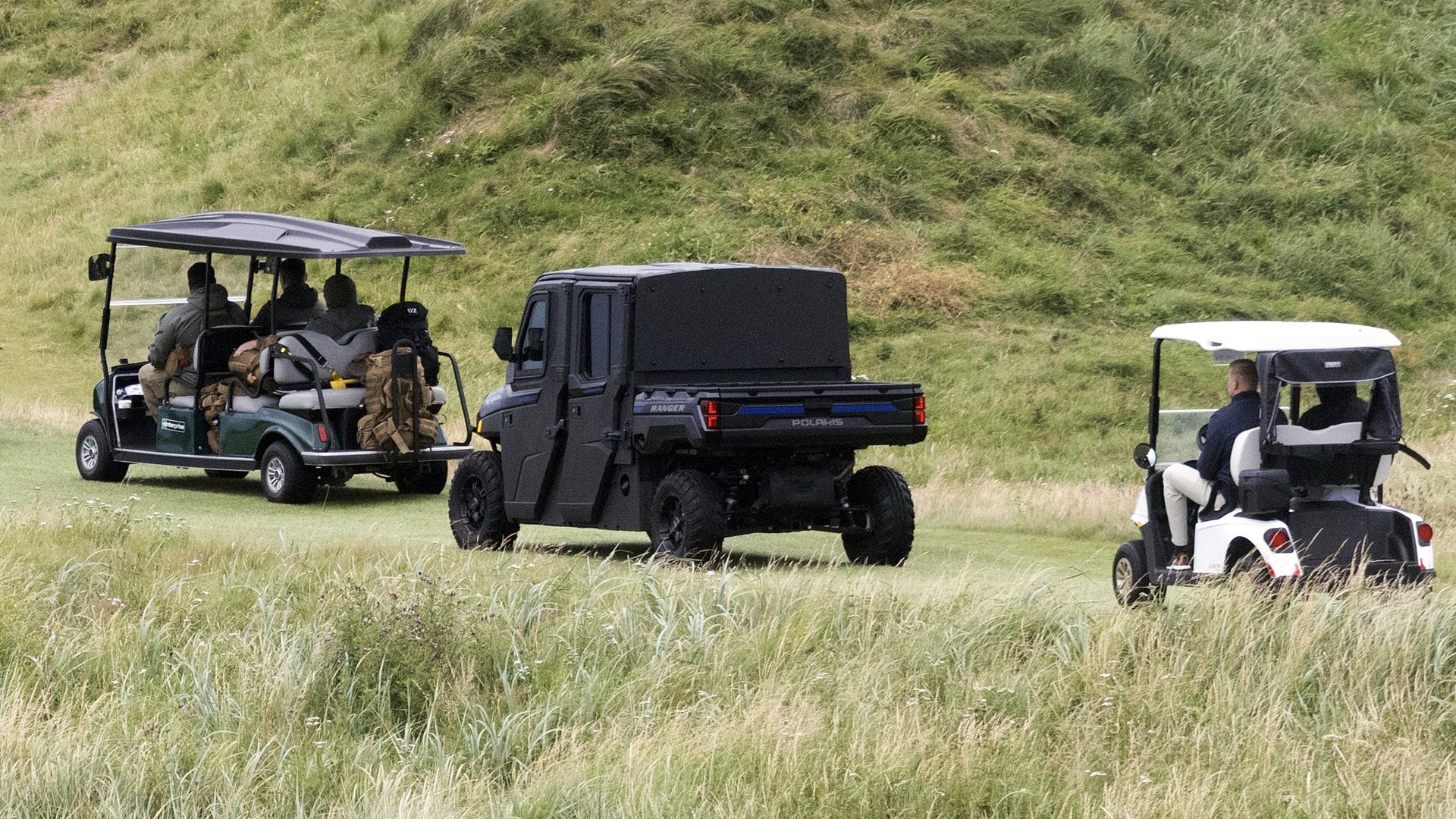 Two regular golf carts and one black fortified golf cart drive on grass near a green hill, with people seated in all three vehicles.