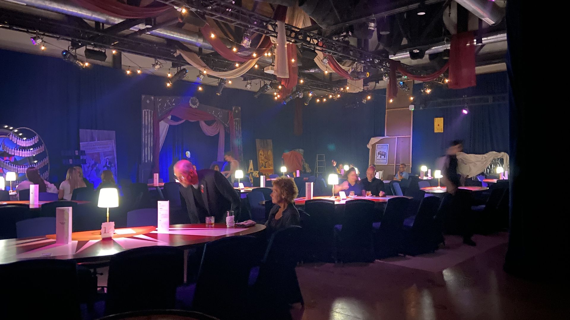 Dimly lit event space with round tables, small white lamps, and people seated or walking; red and white drapes hang from ceiling, creating a cozy atmosphere with blue walls.