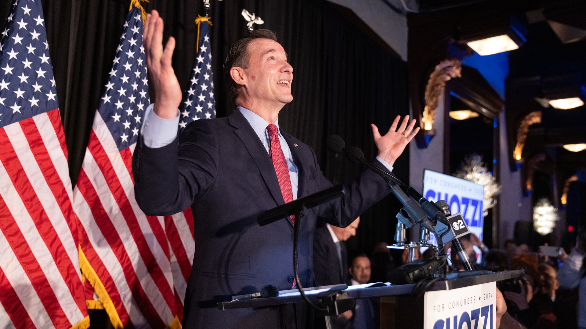 Tom Suozzi, wearing a dark blue suit, light blue shirt and red sign, standing at a lectern in front of American flags.