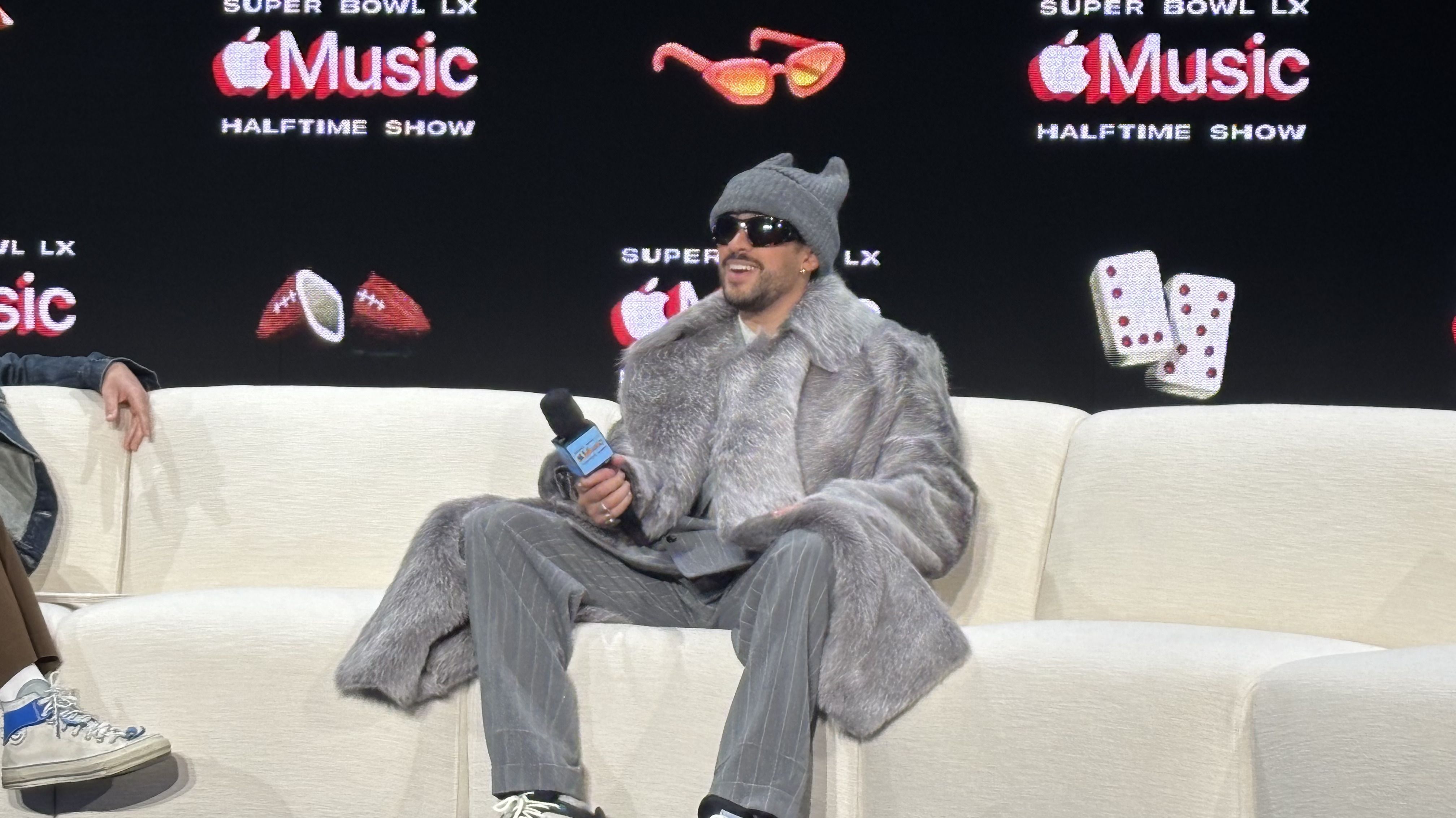 Man in gray fur coat, gray beanie, and sunglasses holding a microphone on a beige couch with a black background featuring "Super Bowl LX Apple Music Halftime Show" logos.