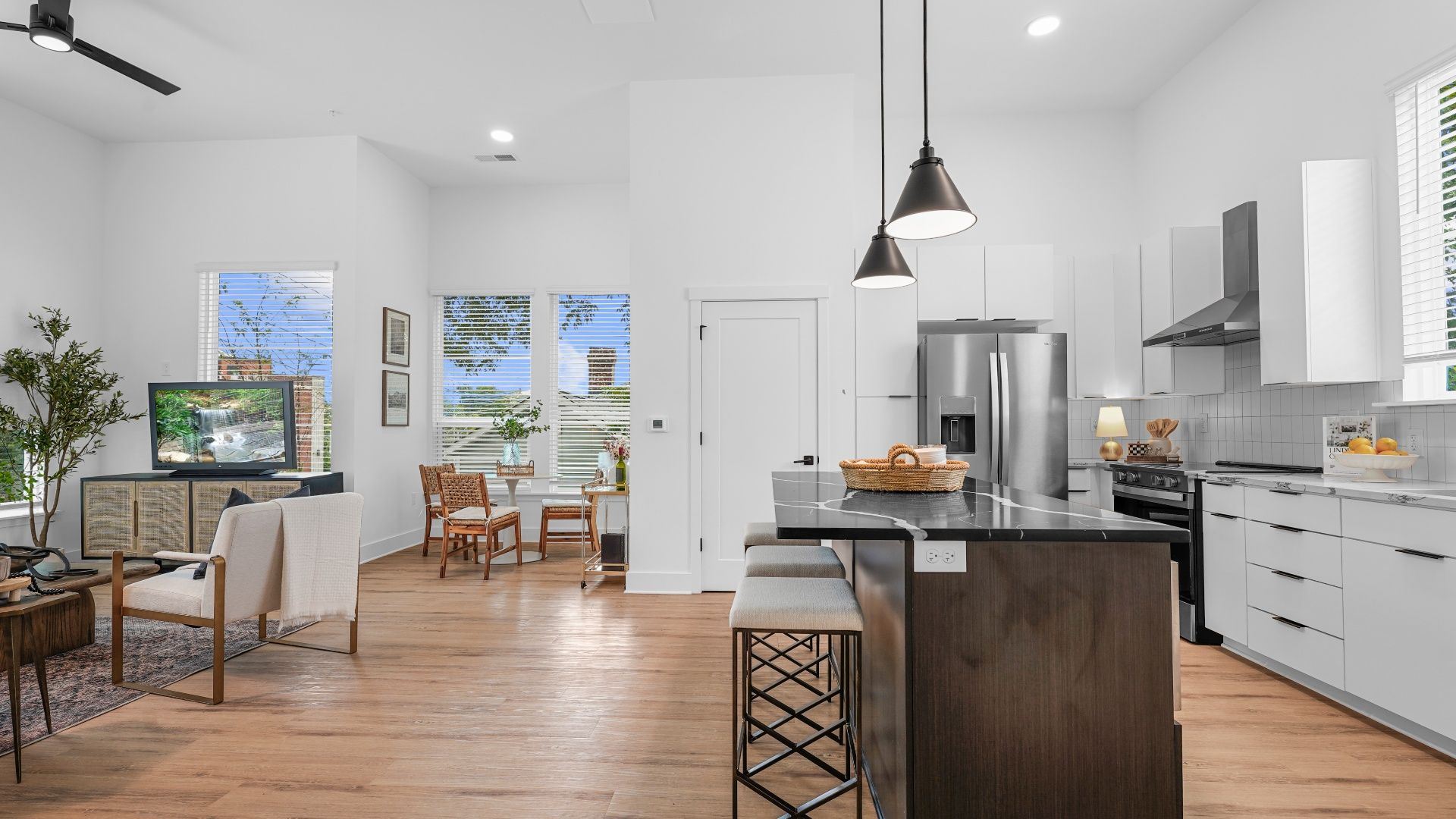 Bright open kitchen and living area with white walls, wooden floor, black kitchen island with stools, stainless steel fridge, wicker chairs, and large windows showing blue sky.