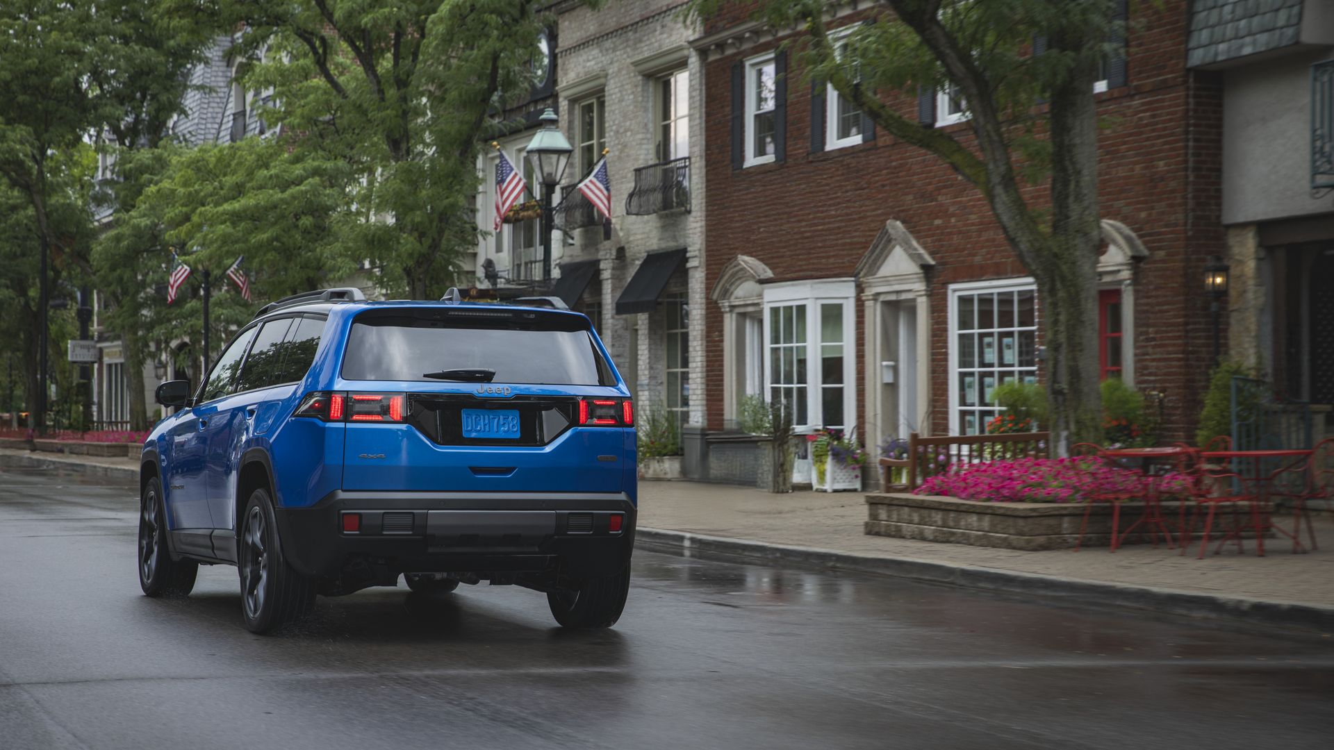 Blue Jeep Cherokee SUV drives down a wet city street lined with brick townhouses, green trees, flag-decorated lampposts, and pink flowers with red outdoor chairs on the sidewalk.