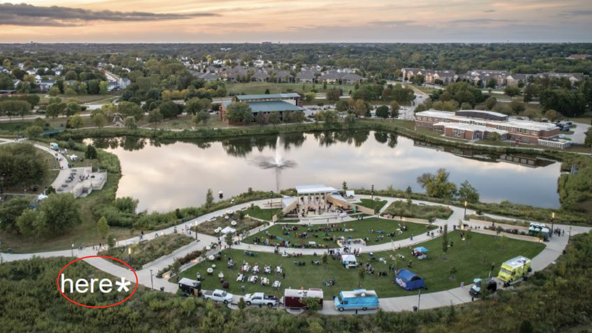 Aerial view of a lakeside park at sunset: circular lawn with a stage and audience, tents, food trucks, and parked cars; a fountain in the lake; trees and distant buildings; "here" circled bottom-left.