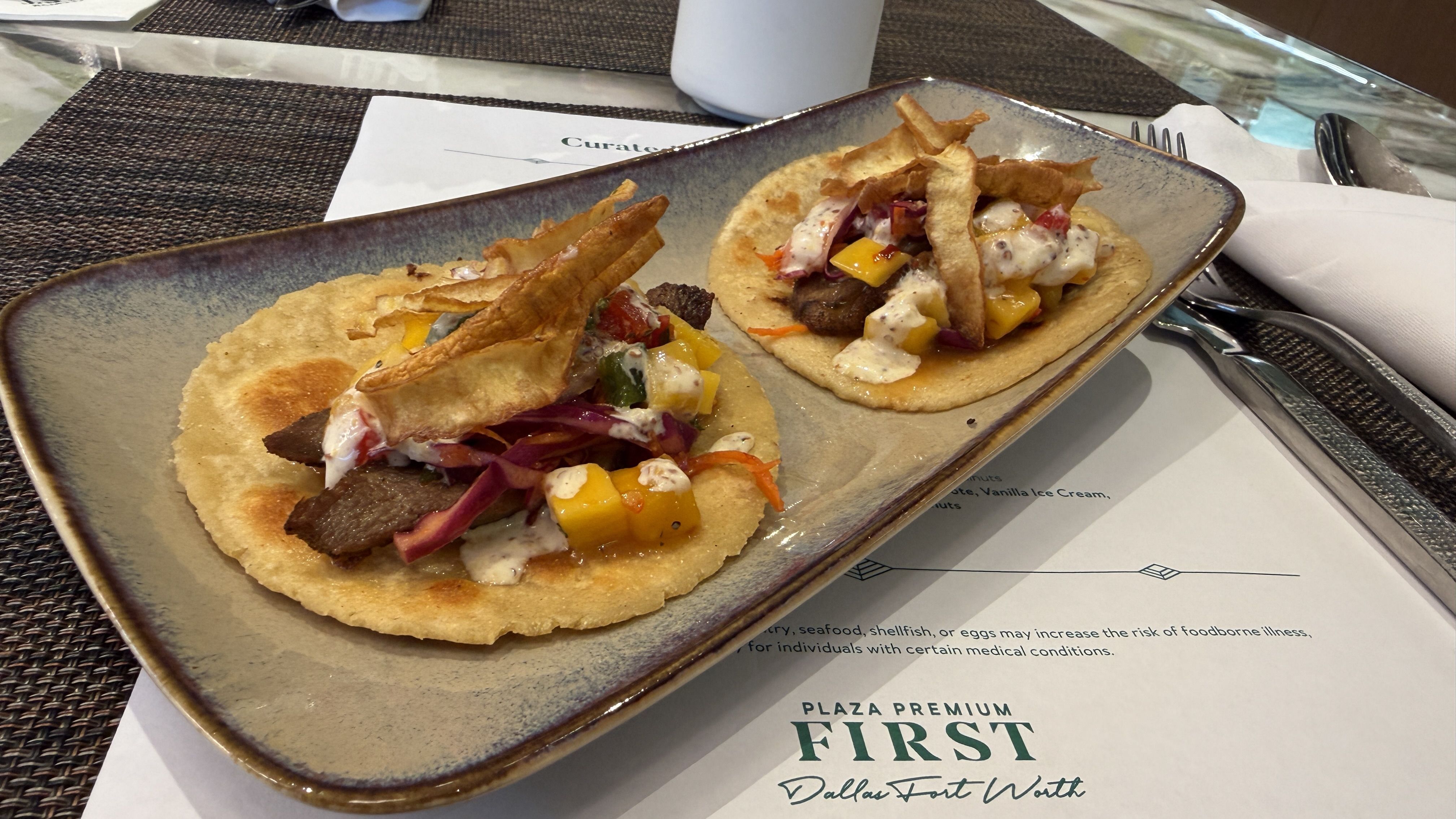 Two beef tacos on a rectangular ceramic plate, topped with mango, slaw, crema, and crispy tortilla strips; placed on a woven placemat with a napkin and silverware nearby.