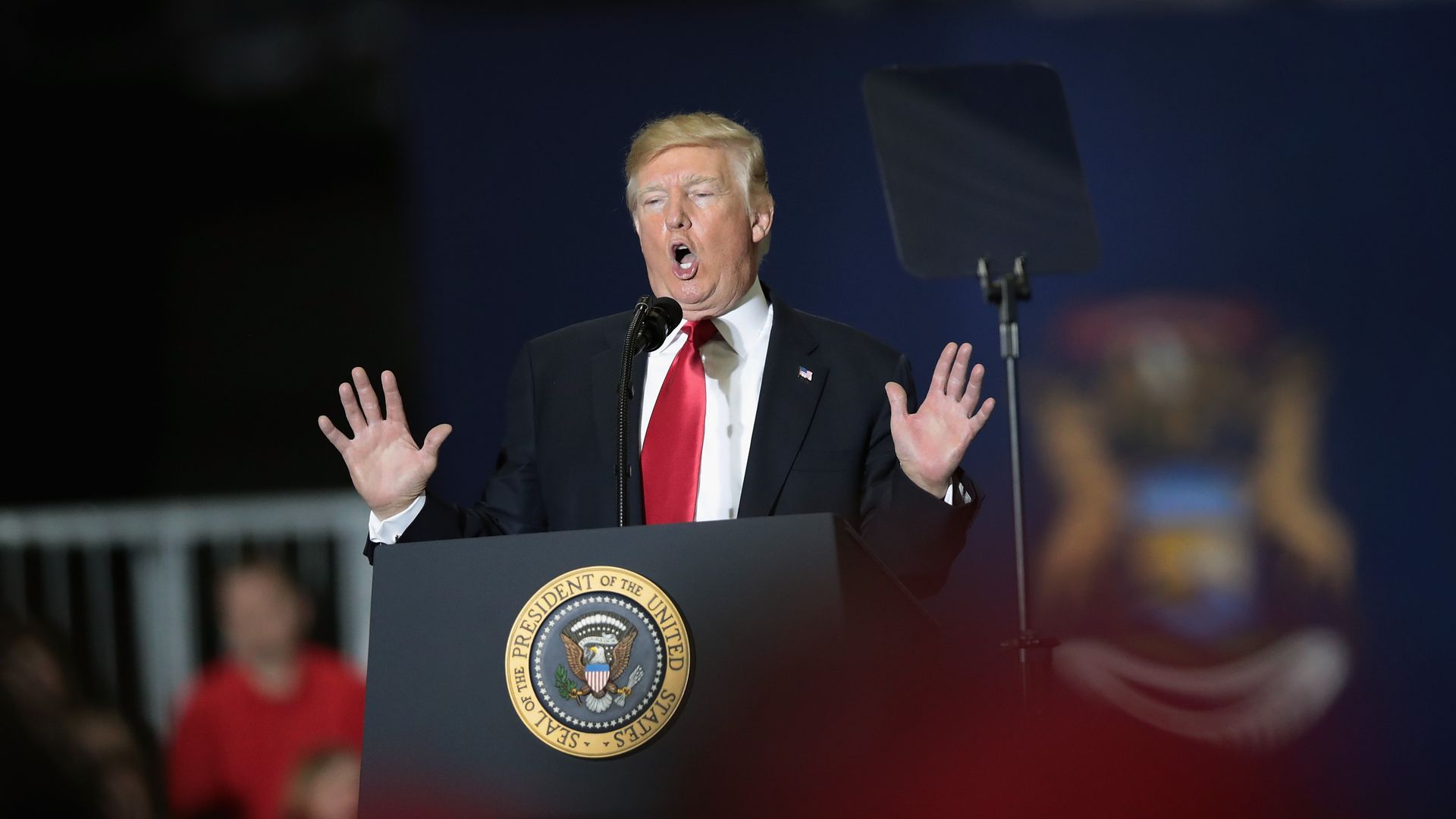 Donald Trump at a podium with a red tie. His hands are up and open to his sides. His mouth is open. 
