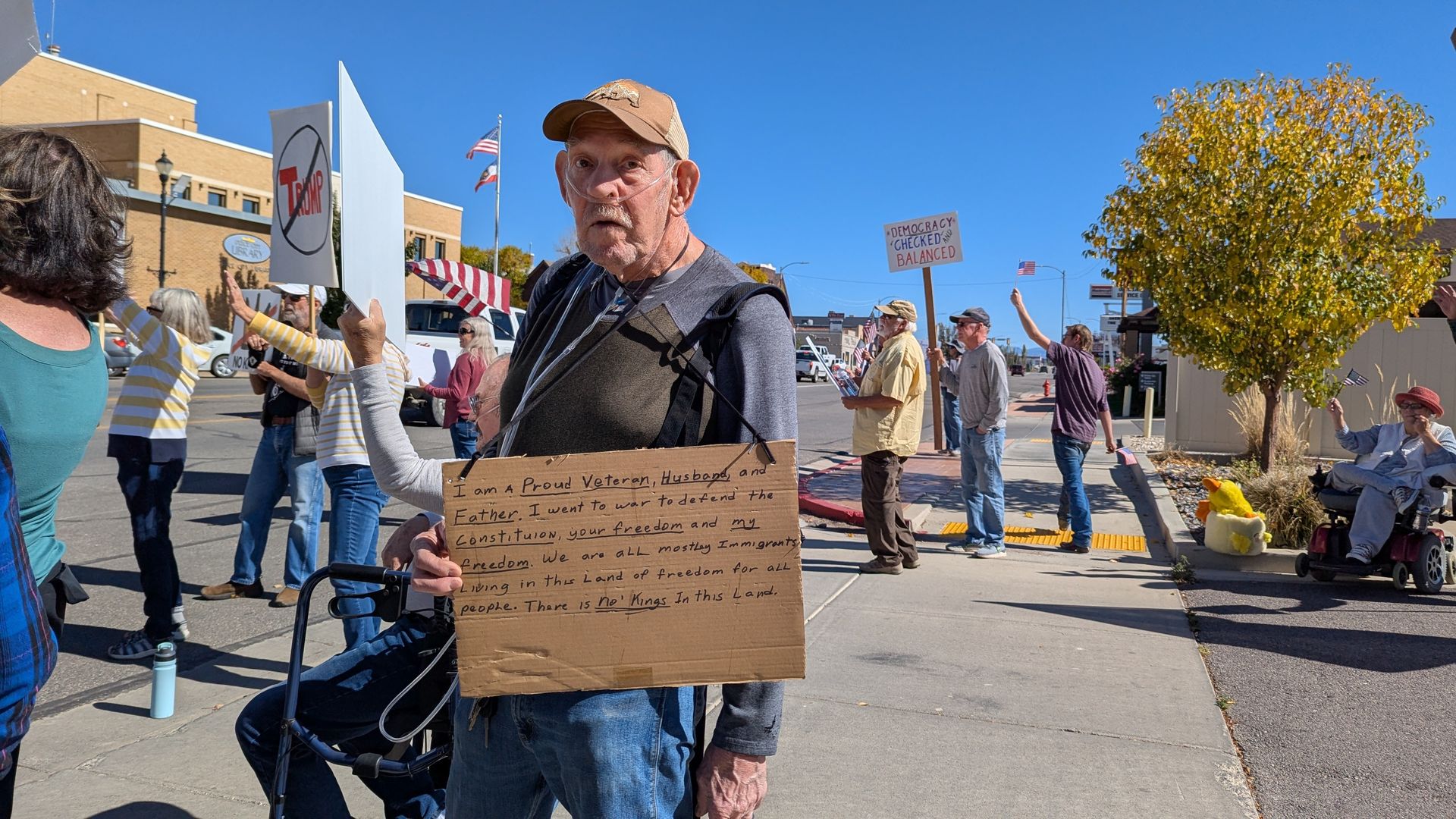 A protester on a small town street holds a cardboard sign that states: "I am a proud veteran, husband and father. I went to war to defend the Constitution, your freedom and my freedom. We are all mostly immigrants living in this land of freedom for all people. There is NO KINGS in this land."