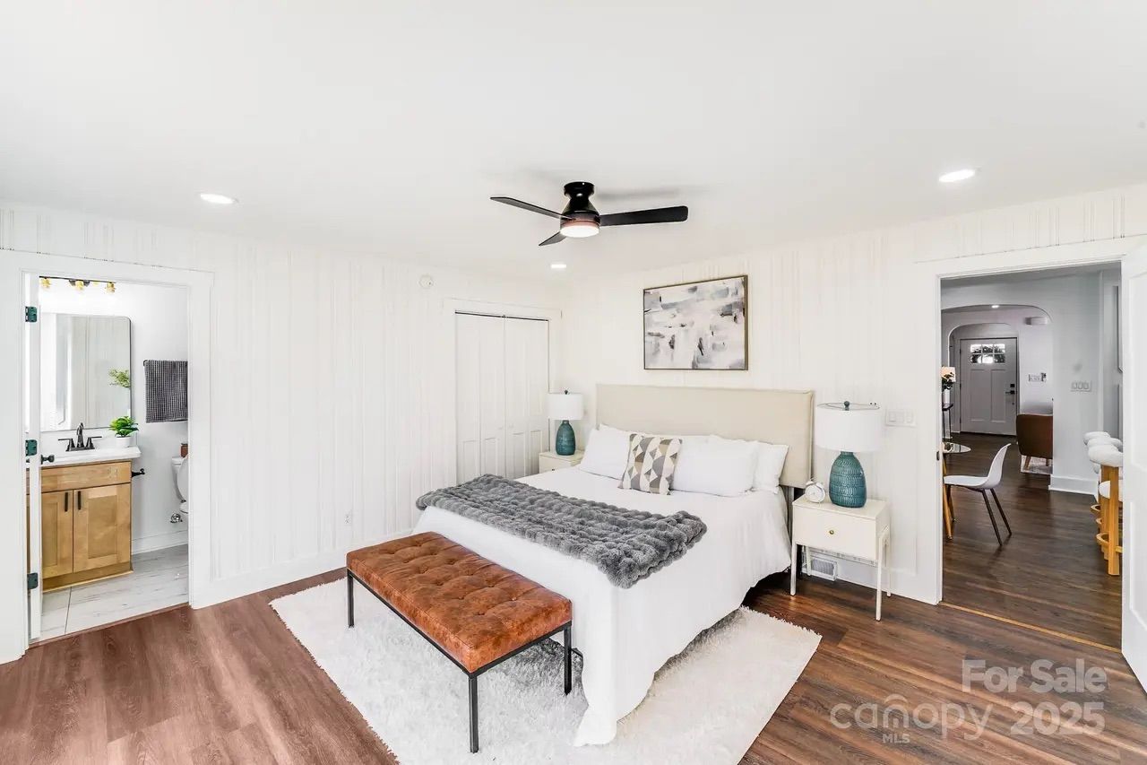 Bright bedroom with white walls, wood flooring, a large bed with white bedding, a gray throw, tan tufted bench, two white nightstands with blue lamps, ceiling fan, bathroom and hallway visible.