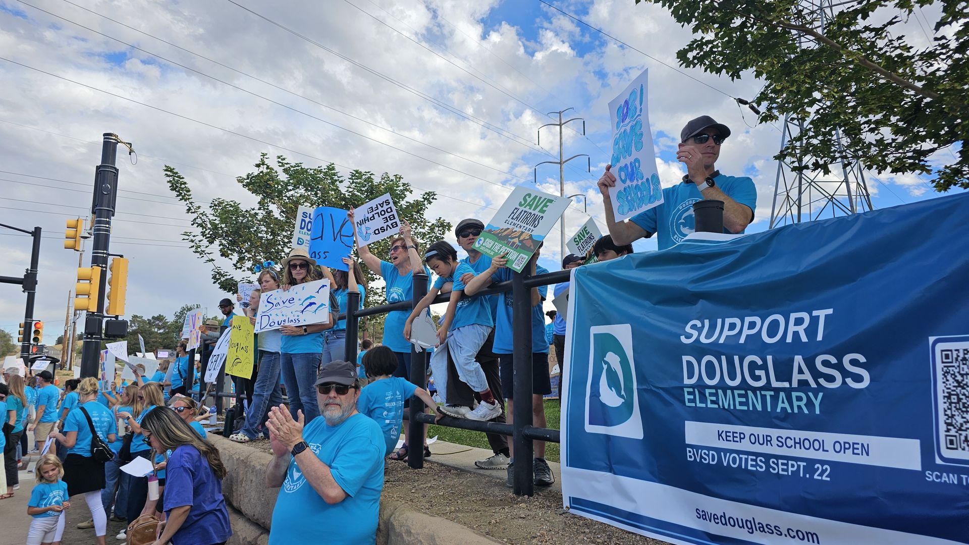 People in blue shirts on a parade float hold signs to save Douglas Elementary; a large blue banner reads "SUPPORT DOUGLASS ELEMENTARY" as power lines and trees loom over a sunny city street.