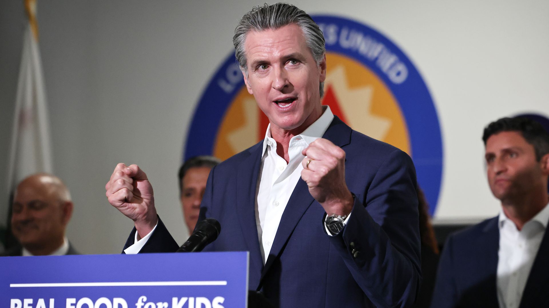 A gray-haired California Gov. Gavin Newsom, wearing a navy jacket and an open white shirt, clenches his fists in front of a sign saying "REAL FOOD for KIDS."