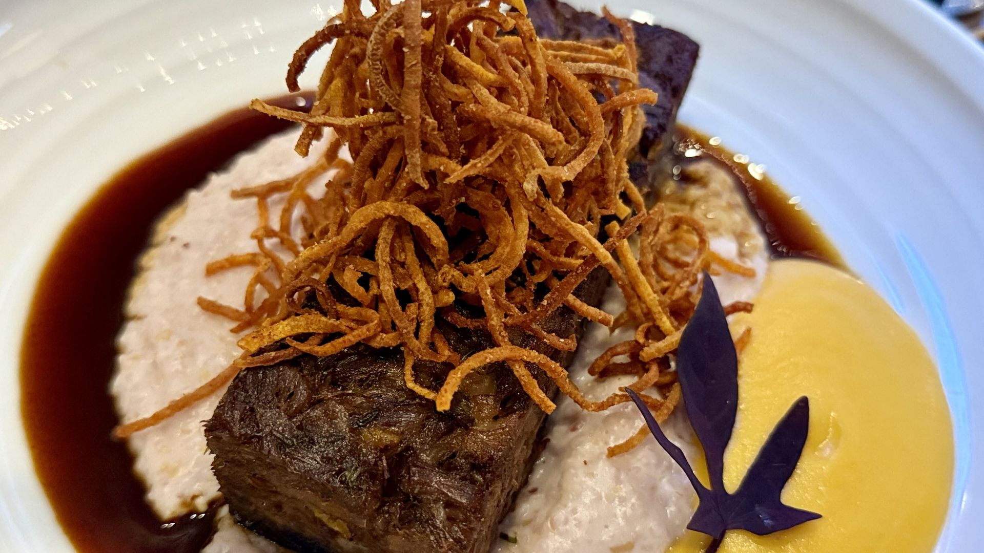 A plated dish with a thick beef cut topped by a nest of crispy shoestring potatoes, brown sauce, creamy mashed potatoes, a yellow sauce, and a purple edible flower garnish.