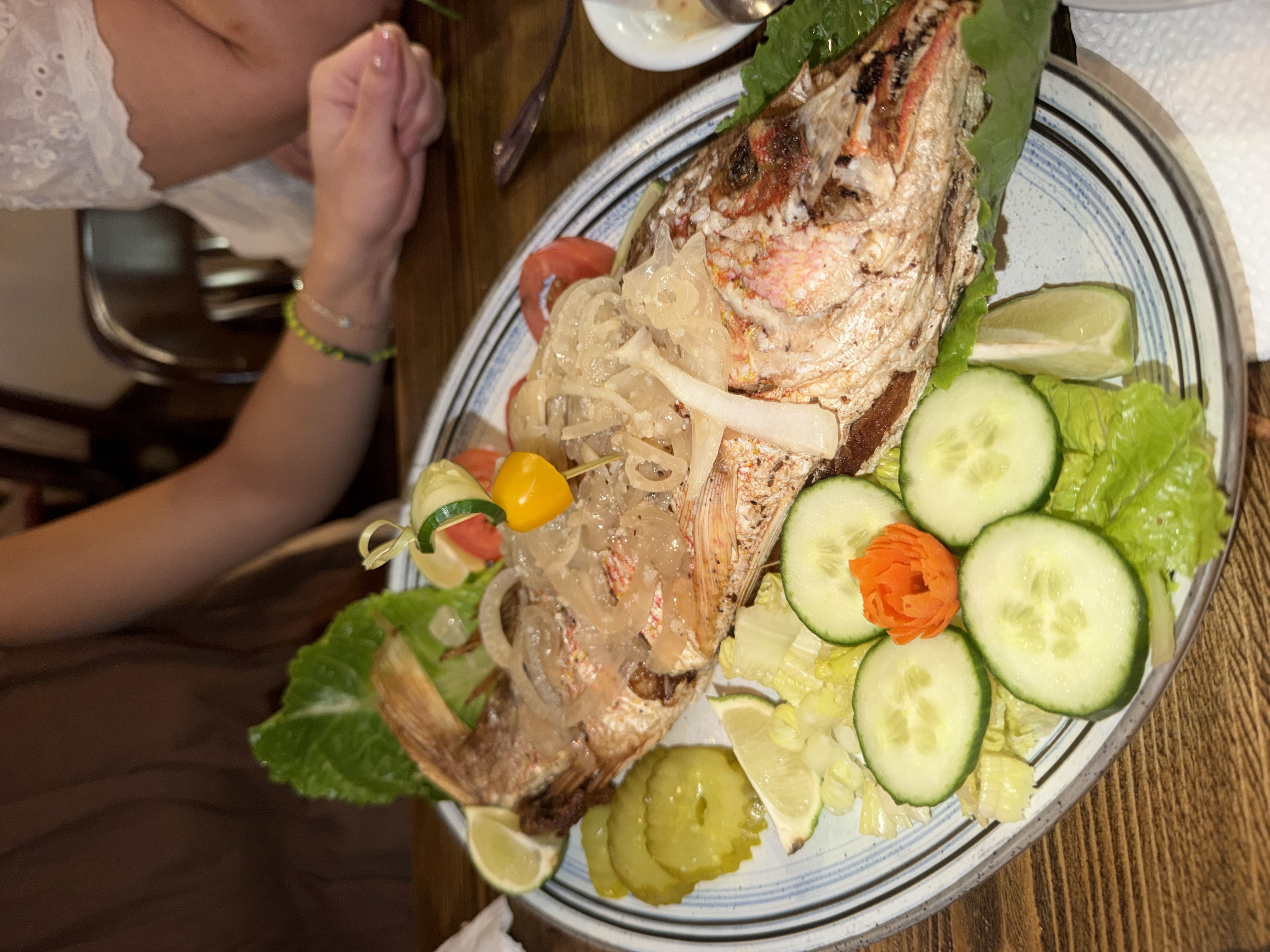 Whole grilled fish on a plate, surrounded by sliced cucumbers, onions, lettuce, lime wedges, and a decorative pepper skewer; a person in lace sleeves sits at the table nearby.