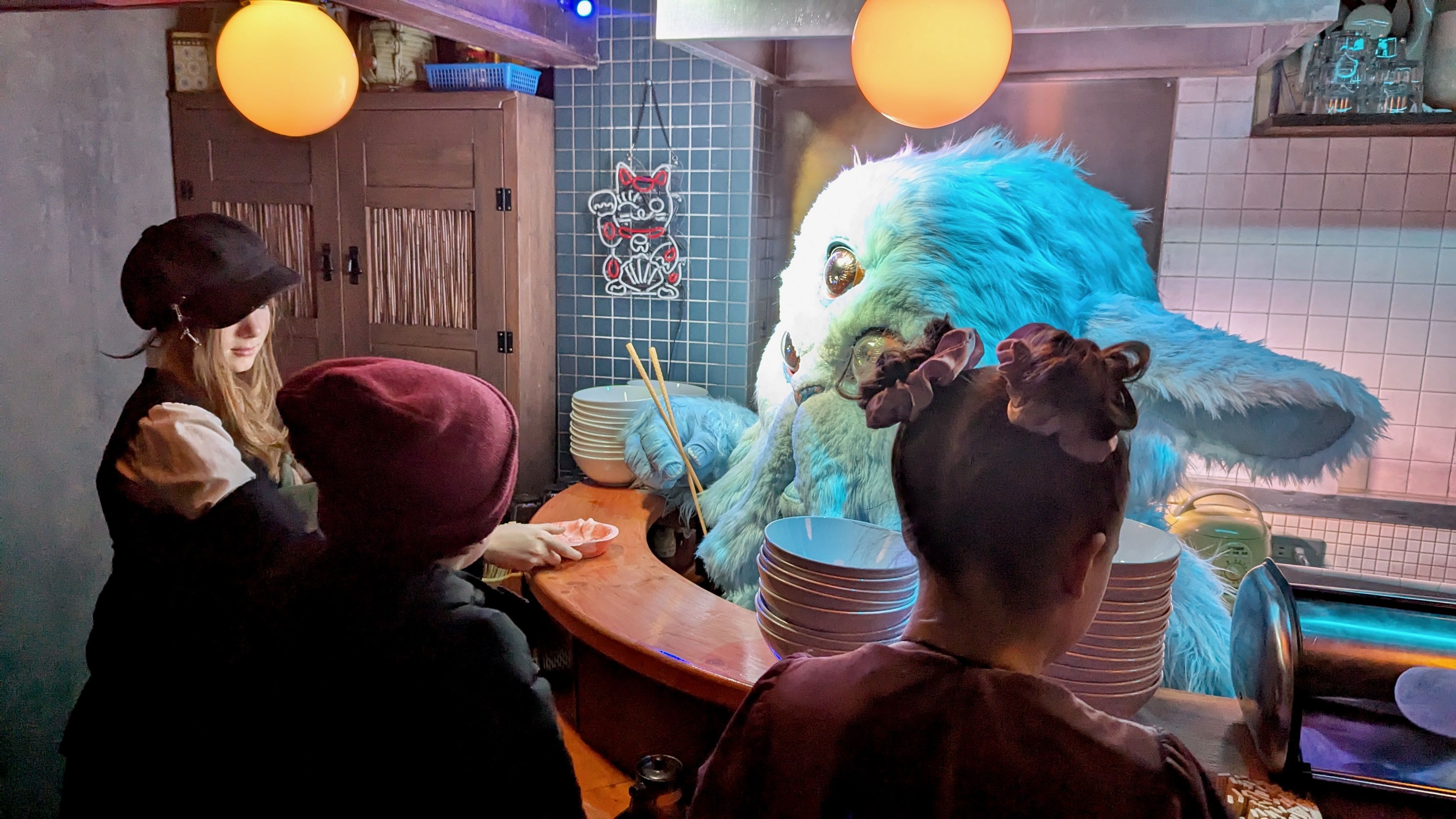 Three kids surround a fluffy animatronic at what looks like a ramen bar.