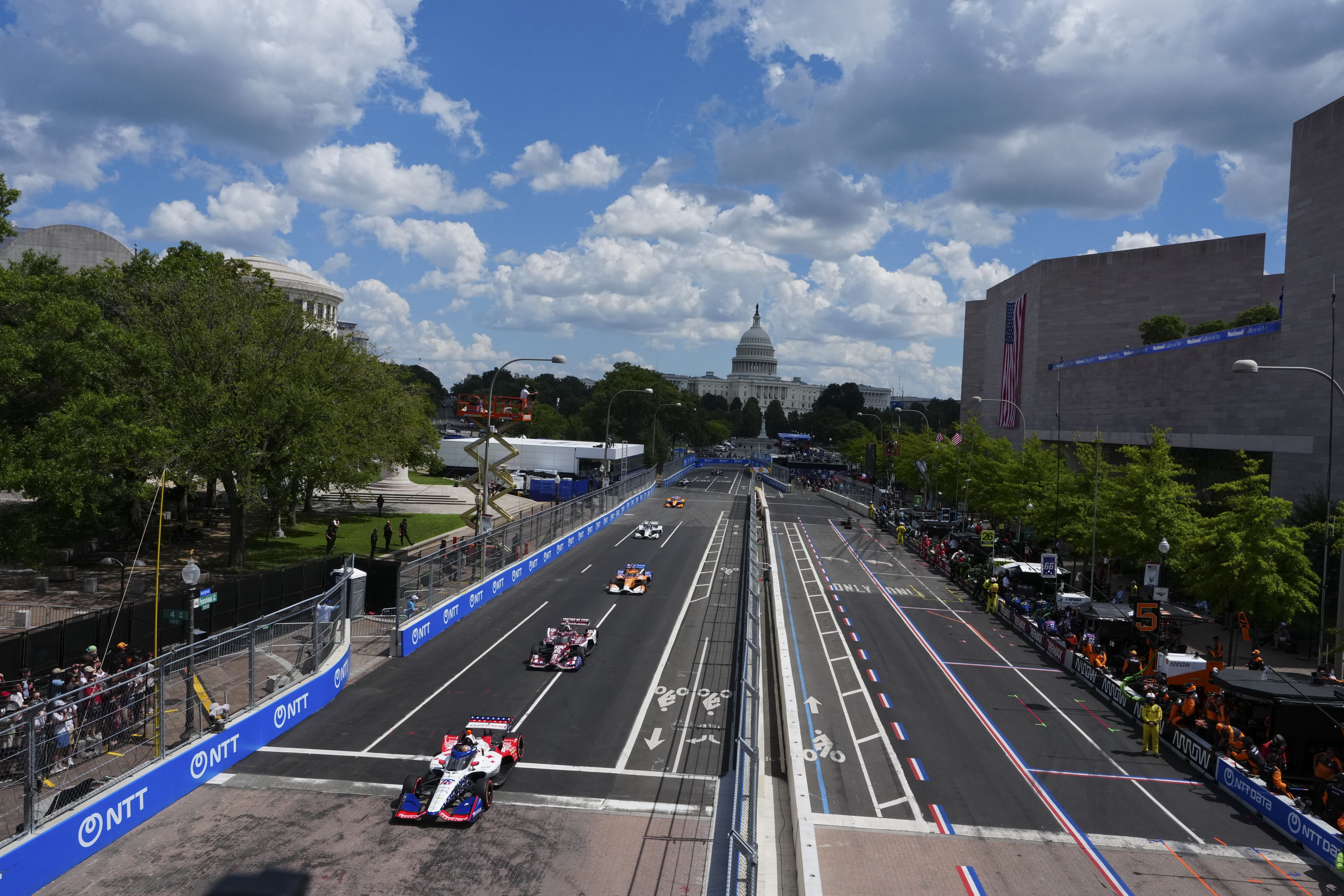 Wide city street race in Washington, DC: race cars on a closed road with blue barriers, green trees on the left, a large gray building and US flag on the right, and the Capitol dome in the distance under a blue, cloudy sky.