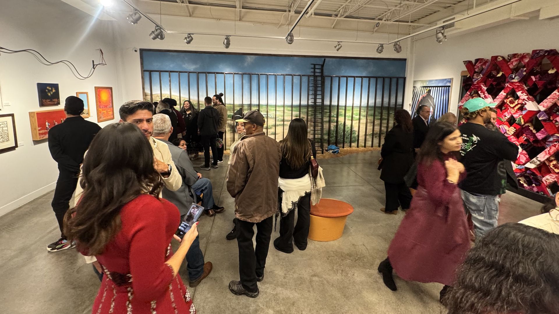 People of diverse ages gather in an art gallery with colorful paintings and a large mural of a barred fence overlooking a green landscape under a blue sky.