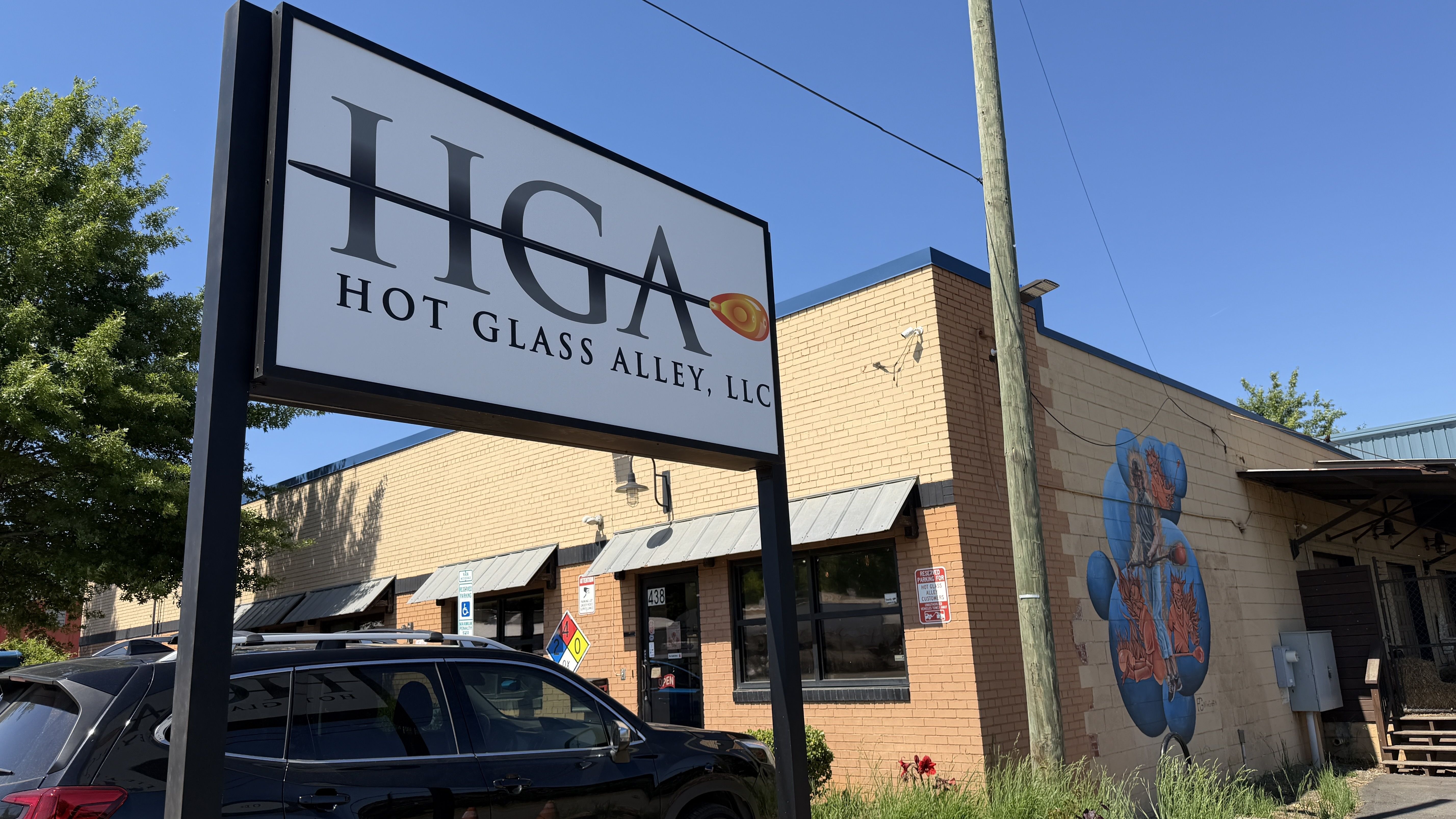 Outdoor scene with a white sign reading "HGA Hot Glass Alley, LLC" and a small orange glass piece, mounted beside a beige brick building with a mural on the wall, blue sky, and a utility pole.