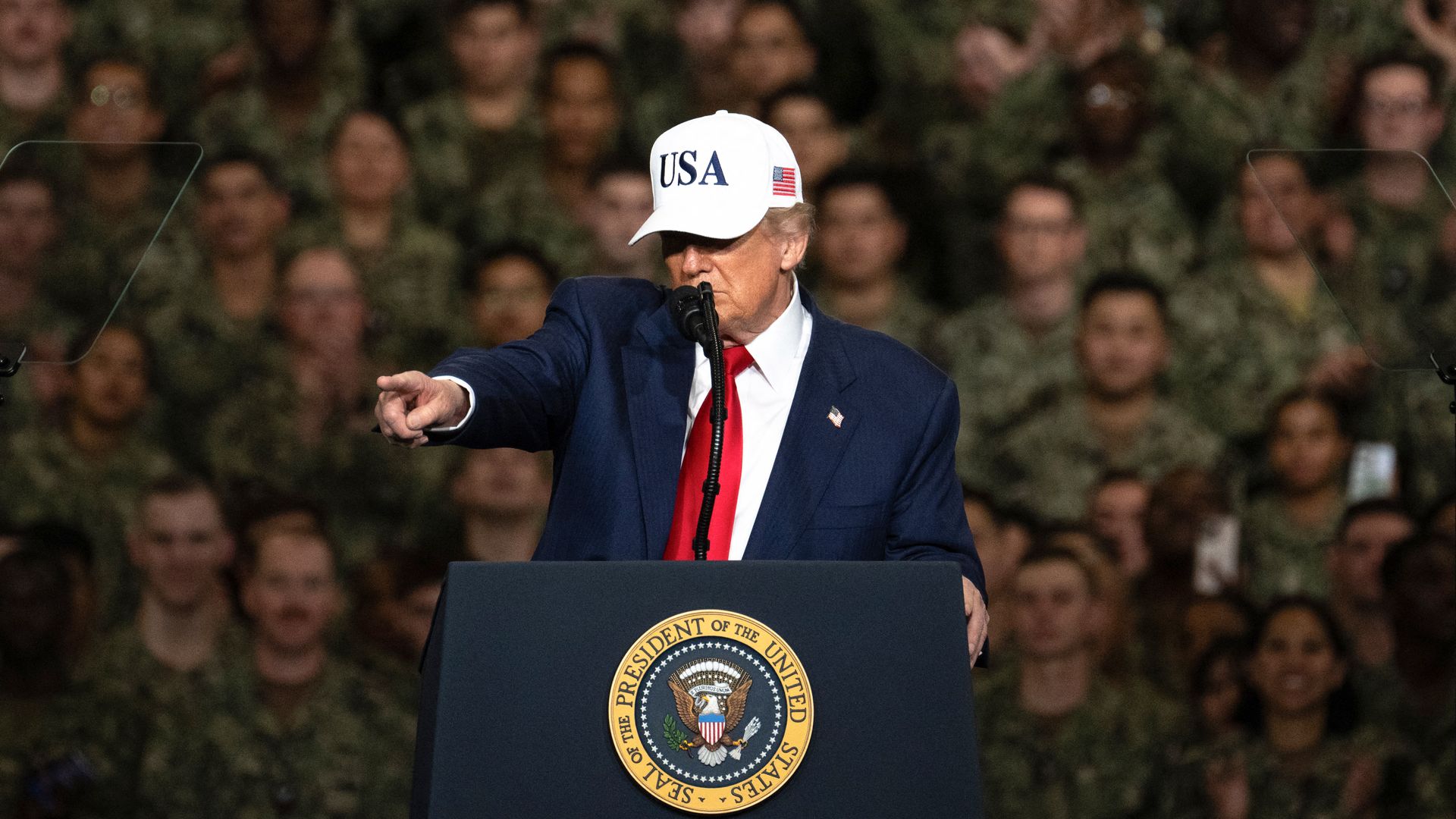 Trump points as he speaks from behind a lectern with a crowd of troops visible behind him. He's wearing a blue suit, white shirt, red tie and white hat that reads "USA" on the front. 