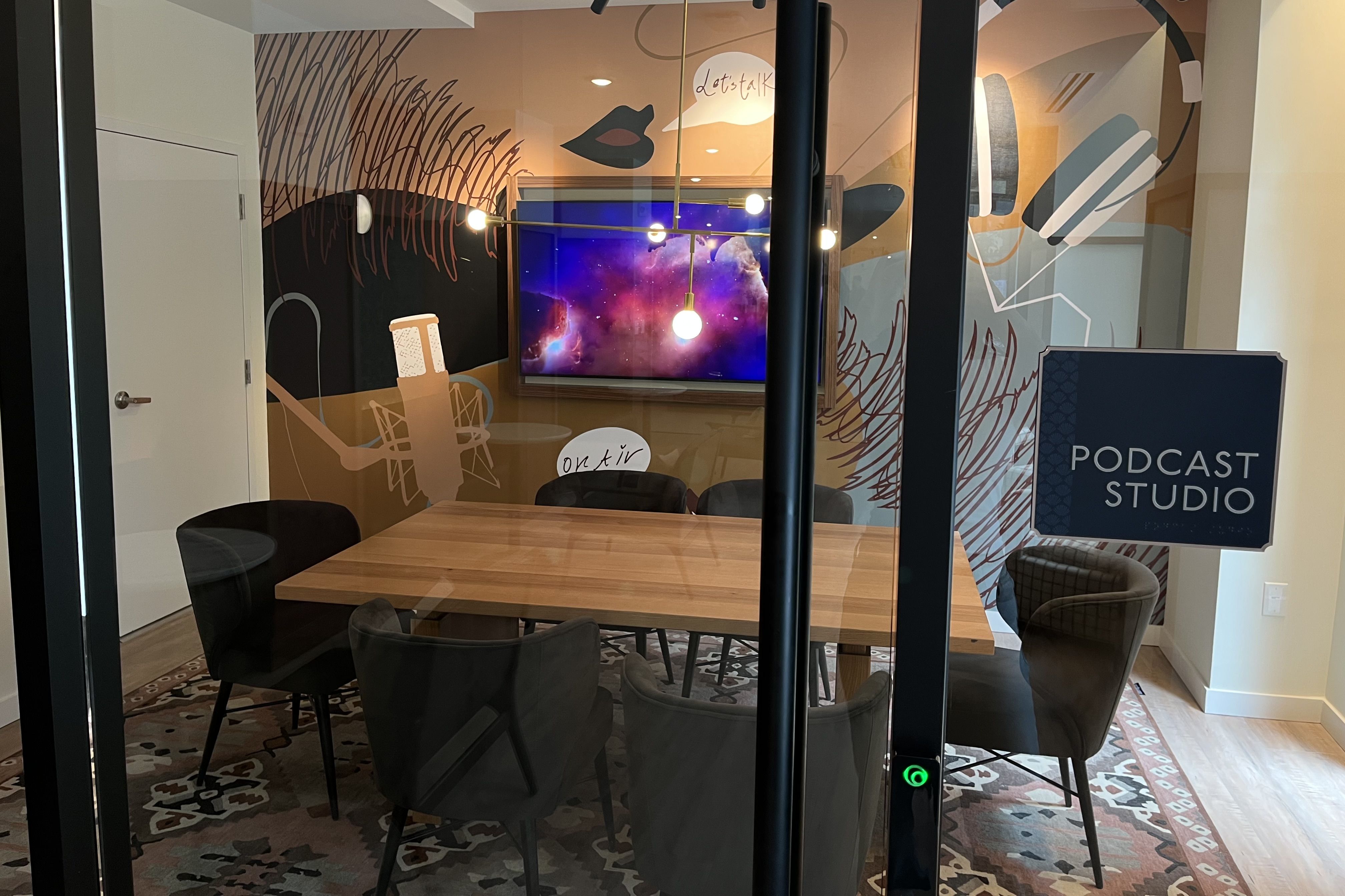 Podcast studio with glass walls, wood table, black chairs, a colorful space-themed screen, and wall art of microphone, lips, headphones, and speech bubbles saying "Let's talk" and "on air".