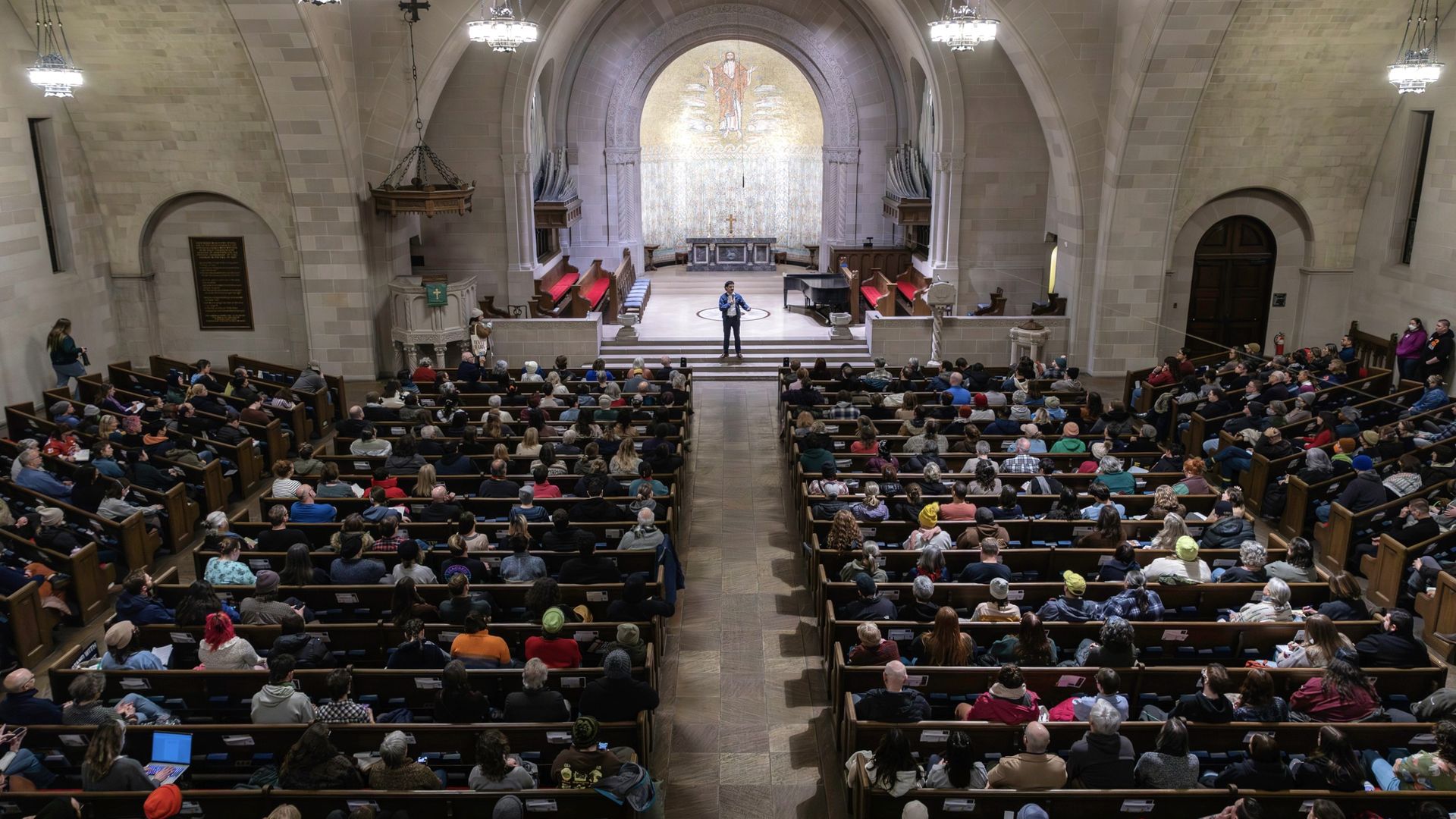 A large crowd seated in pews inside a church or chapel listens to a speaker standing on the altar stage under arched ceilings and chandeliers, with a gold mosaic behind.