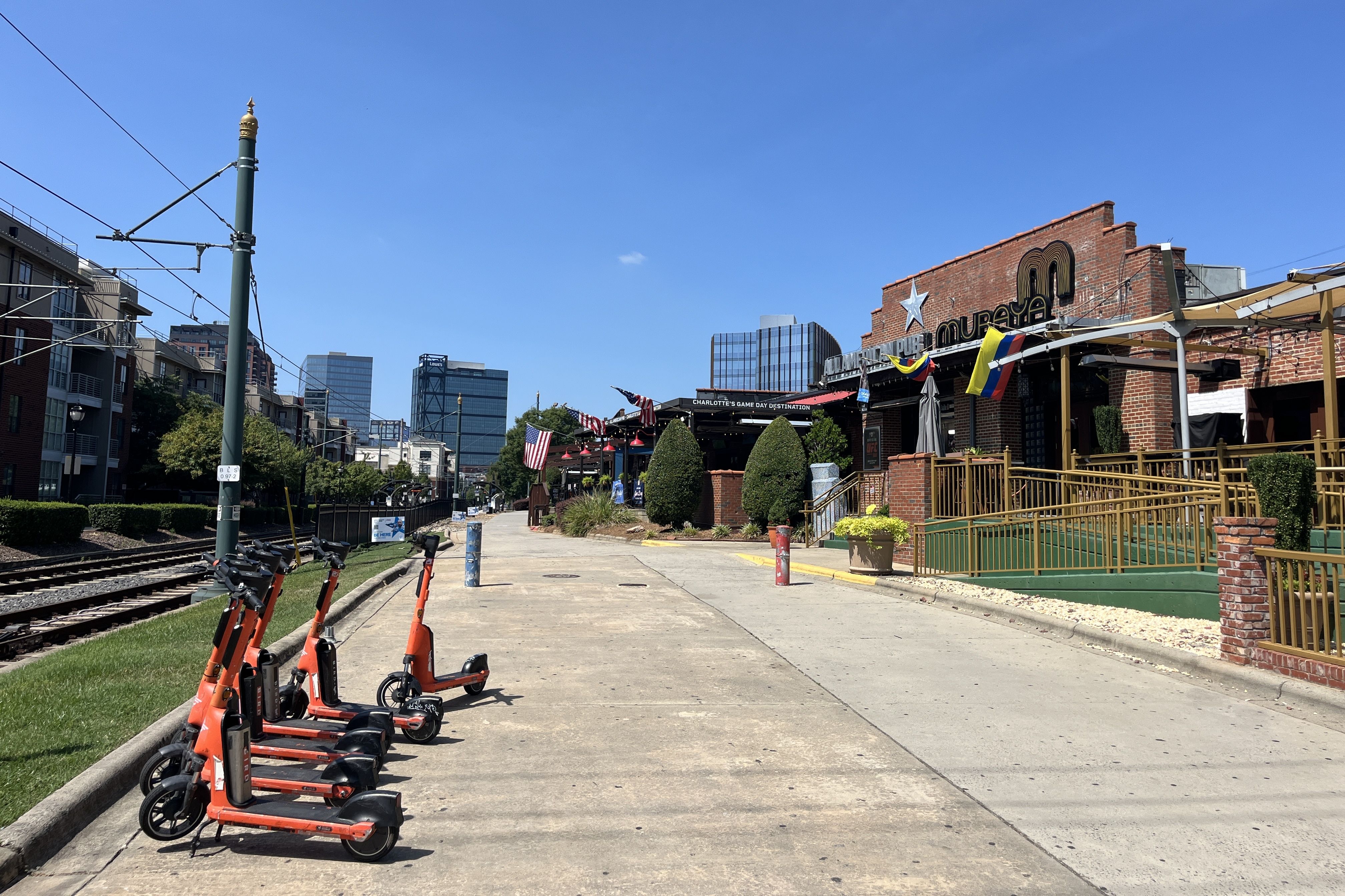 Row of orange rental scooters lined up along a concrete path beside train tracks, with a green pole and overhead wires on the left and a brick building with a logo and flags on the right under a sky.