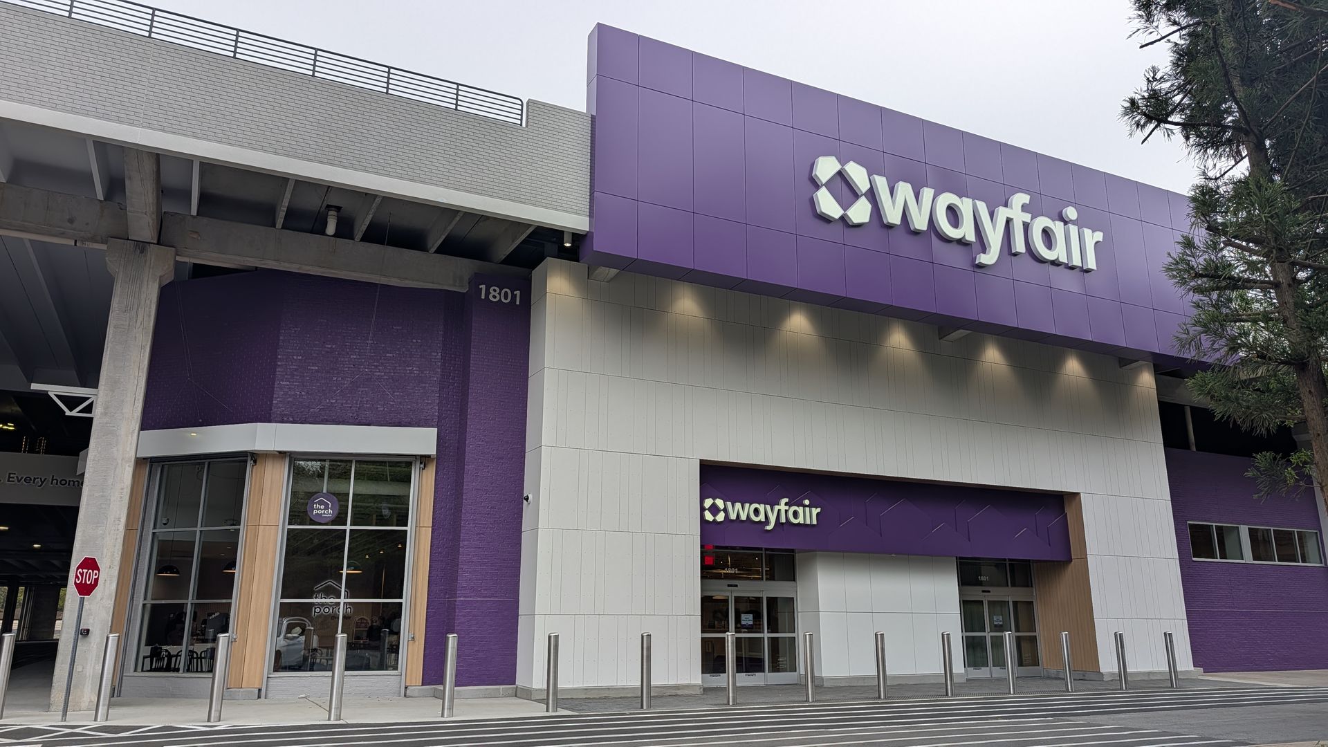 Exterior view of a Wayfair store with a purple facade, white accents, and a large Wayfair logo. Glass entrance, metal bollards, crosswalk markings, a red stop sign, and a tree on the right.