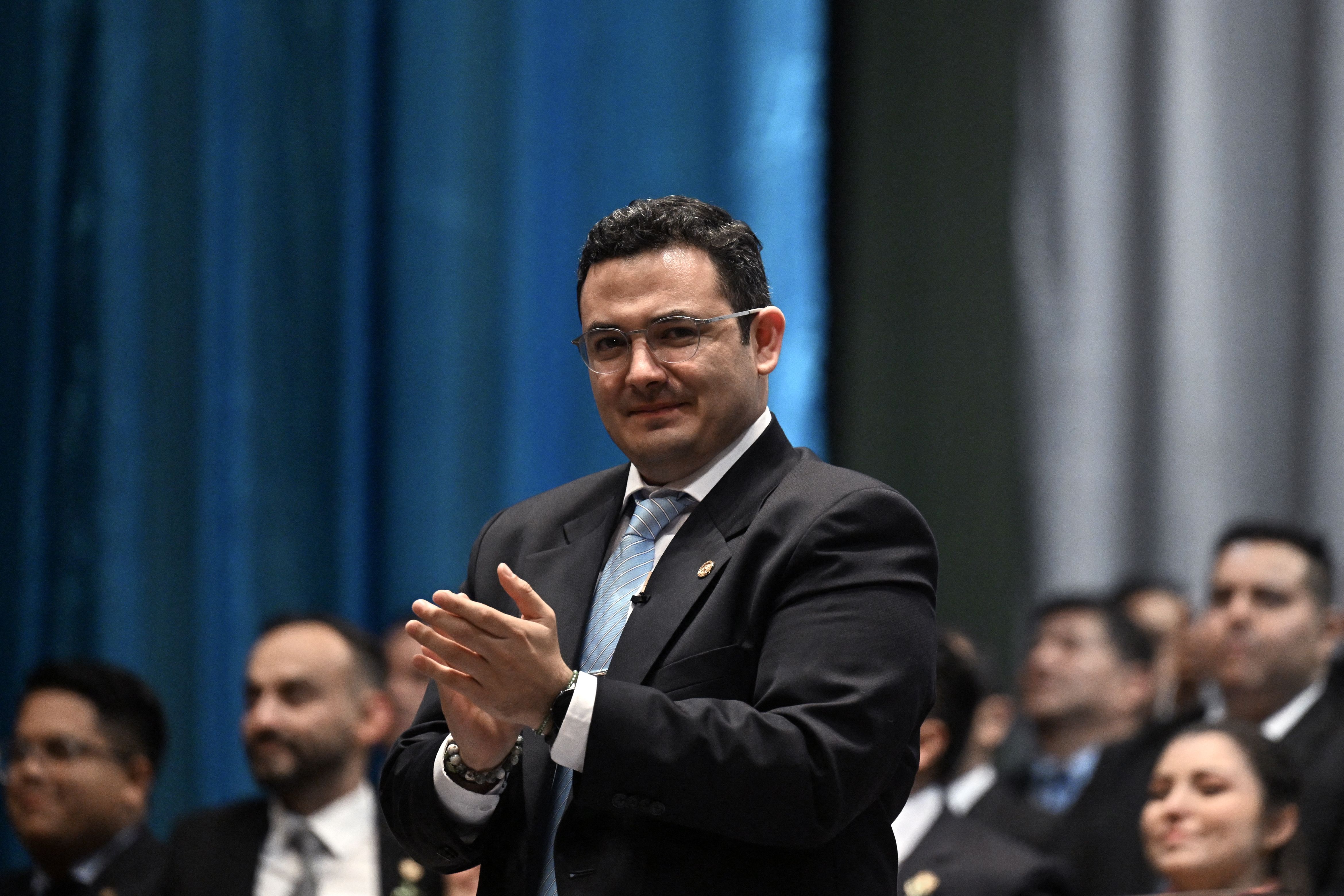 Samuel Perez, the new head of the Guatemalan congress, stands clapping and smiling. He is wearing a baby blue tie, in the color of the Guatemalan flag. 