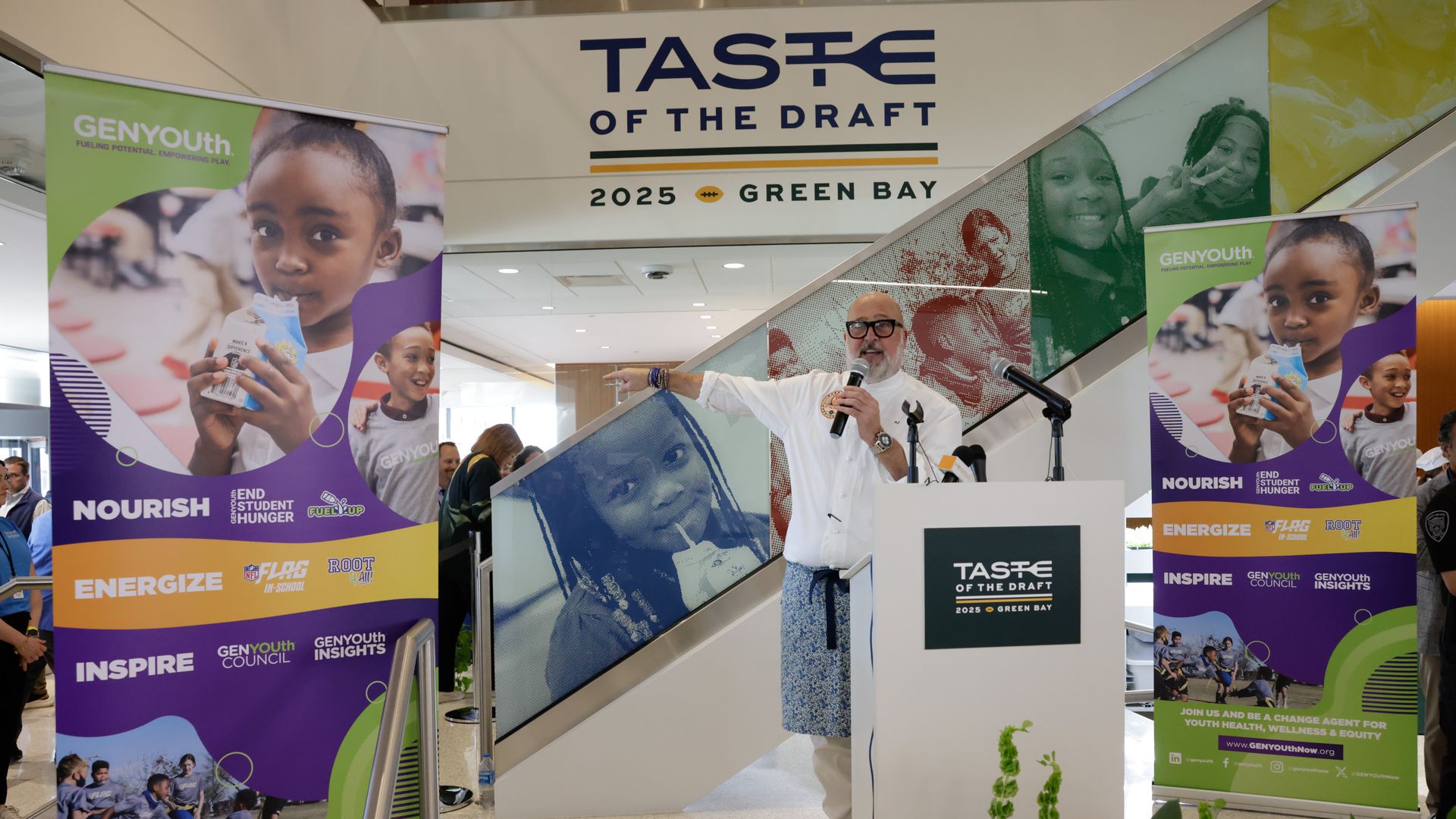 A man with glasses and a white shirt speaks into a microphone at a podium marked "TASTE OF THE DRAFT 2025 GREEN BAY." Bright banners with children and GENYOUTH flank the scene.