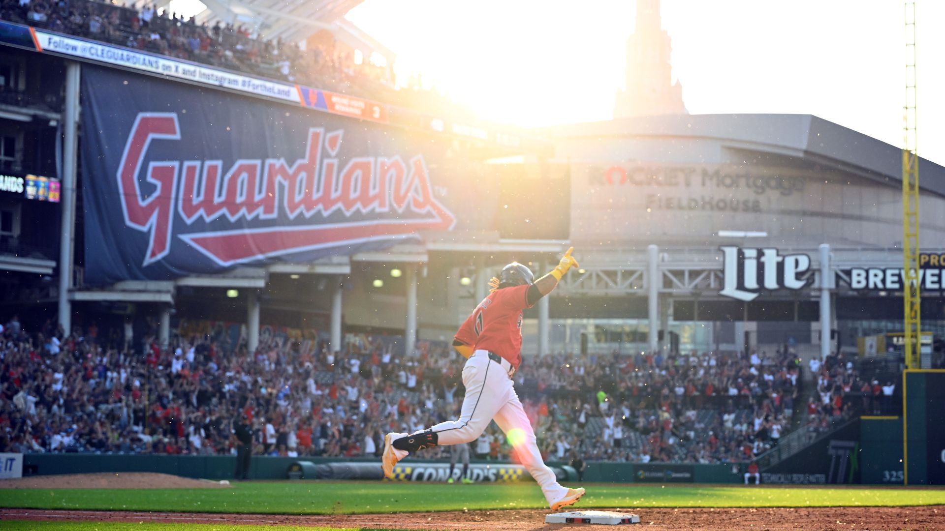 Baseball player in red jersey runs the bases as sun sets on field with Guardians banner in stands.