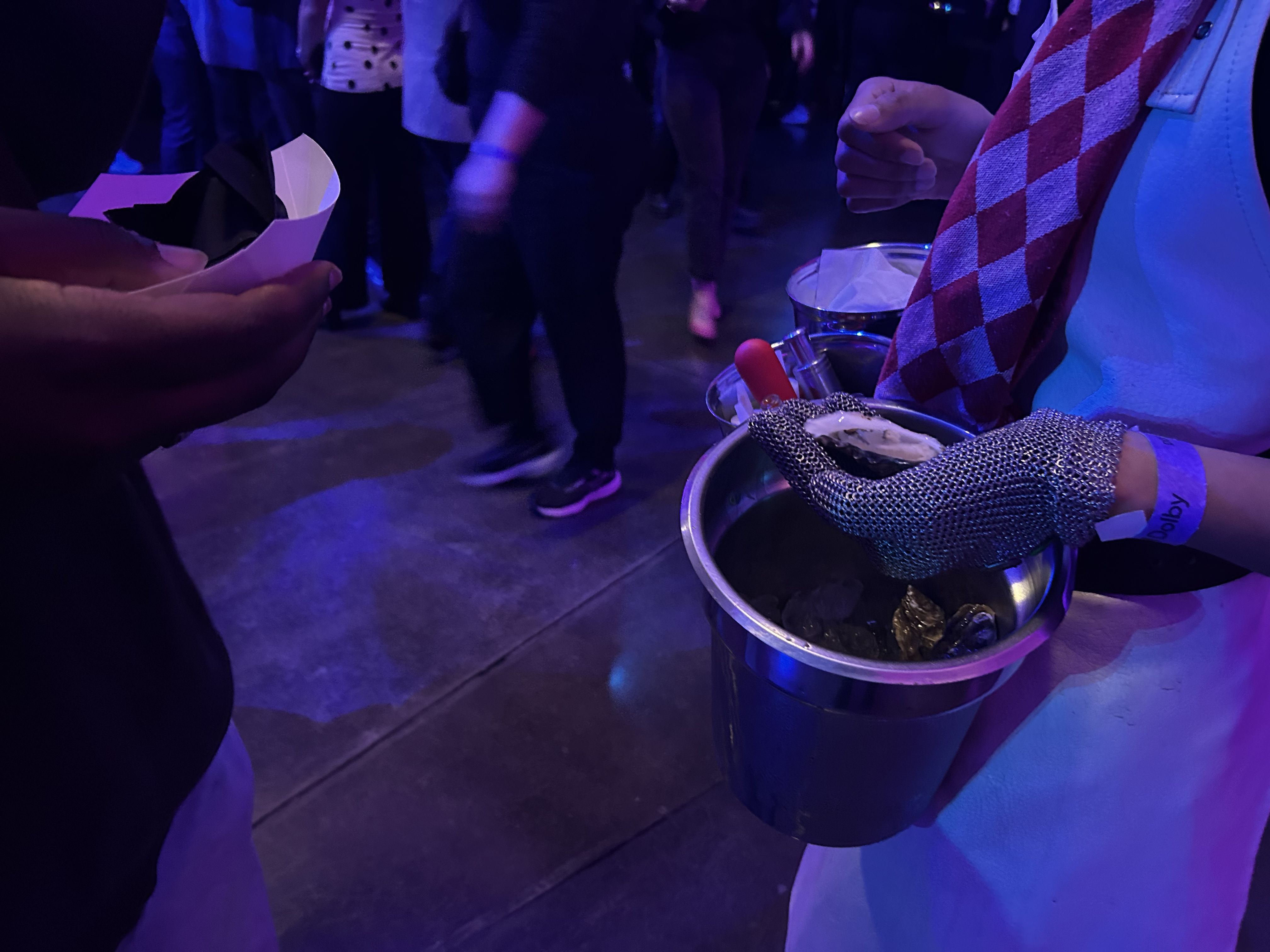 Person wearing a metal mesh glove shucking oysters, holding one oyster over a bucket with ice and shells, with another person holding a food tray nearby, under purple-blue lighting.