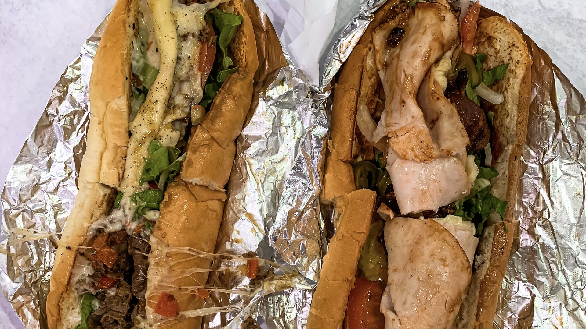 Two sandwiches, stuffed with deli meat and various toppings, sit on pieces of aluminum foil.